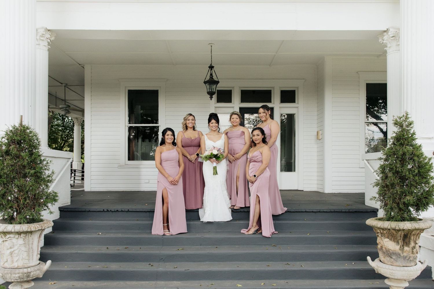 Bridesmaids in pink dresses pose with bride on gray steps of a white building.
