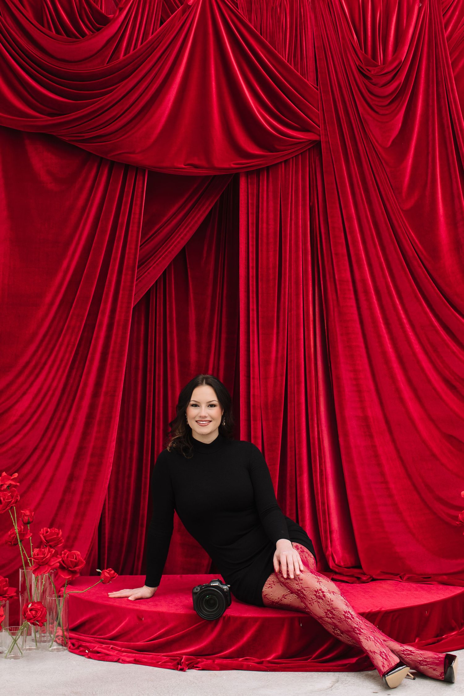 A figure in a black sweater poses against luxurious red velvet curtains with matching red stockings and holiday decor.