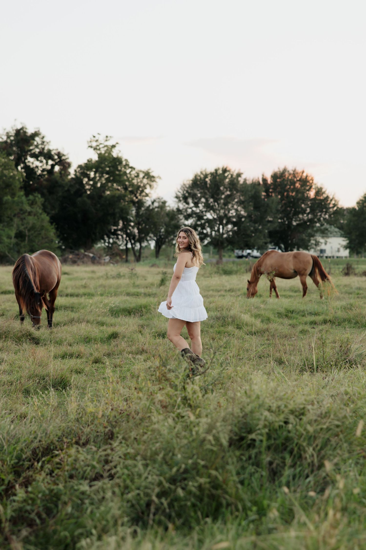 Young woman in white dress and cowboy boots walks through green pasture with two brown horses grazing at sunset.