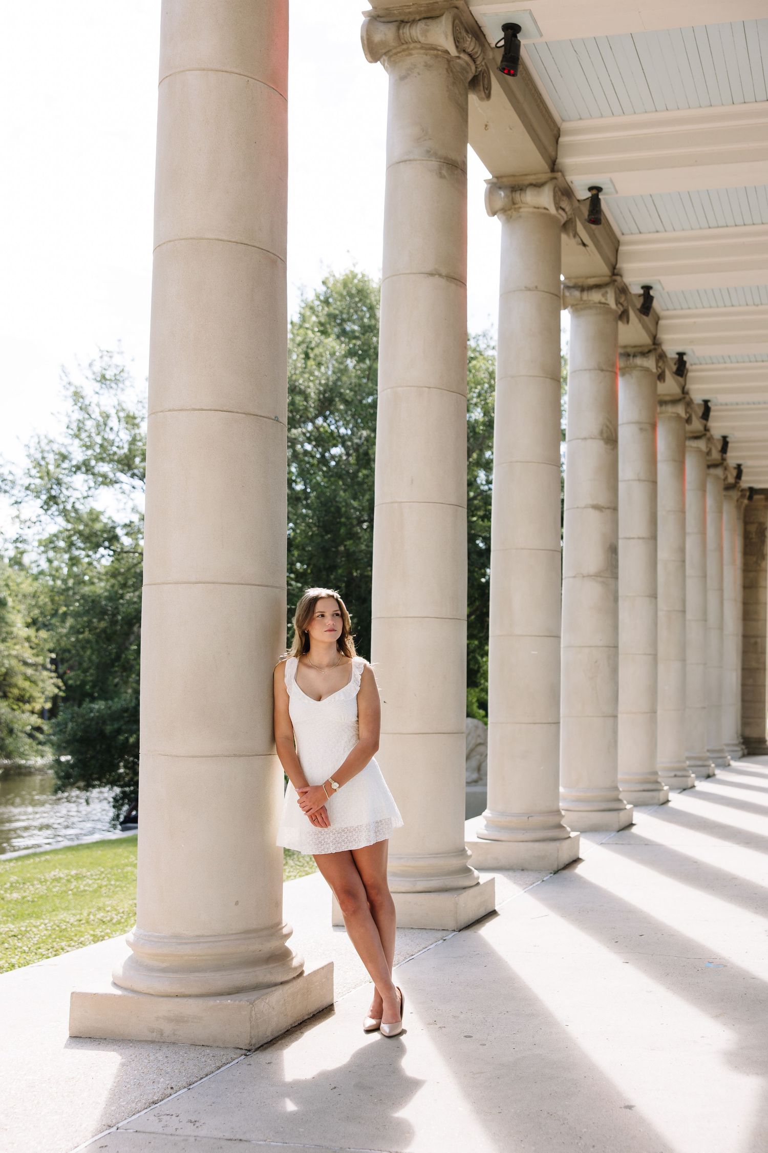 Woman in white mini dress posing between classical columns of elegant colonnade with green trees in background.