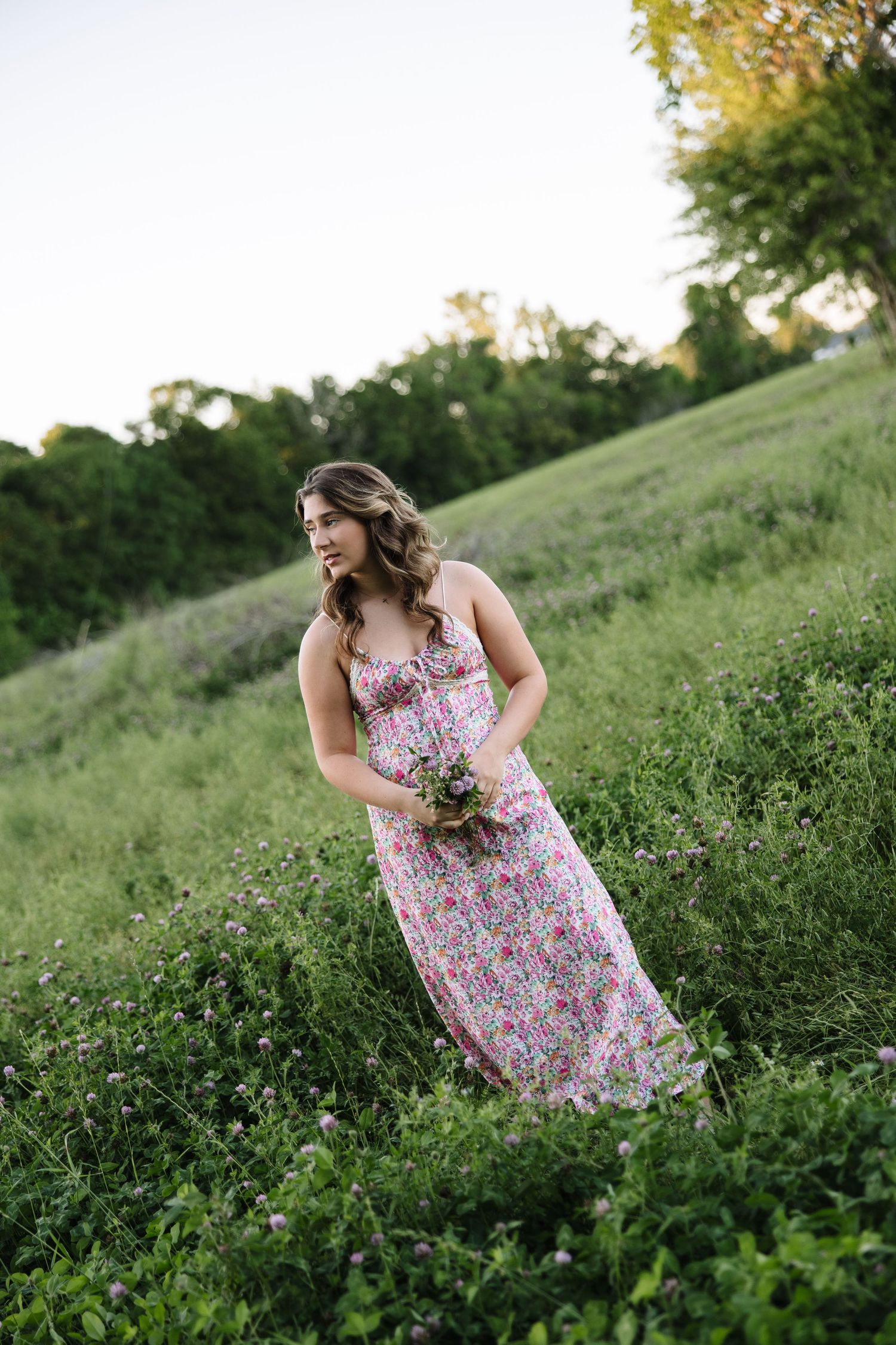 Young woman in floral pink maxi dress holding wildflowers in a lush green meadow at dusk.
