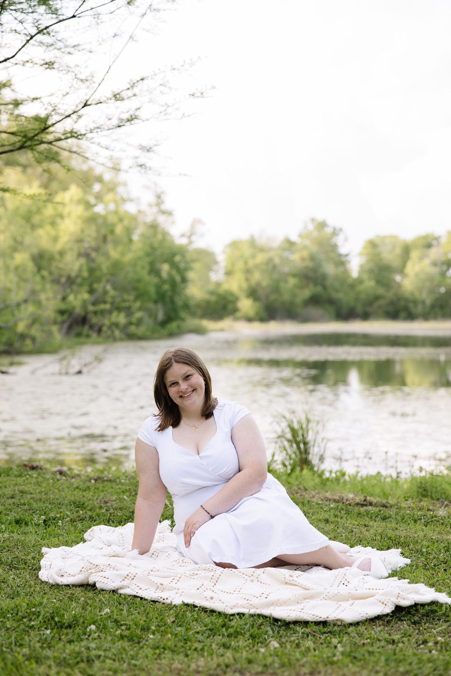 Woman in white dress sitting on blanket by serene pond surrounded by lush green trees on sunny day.