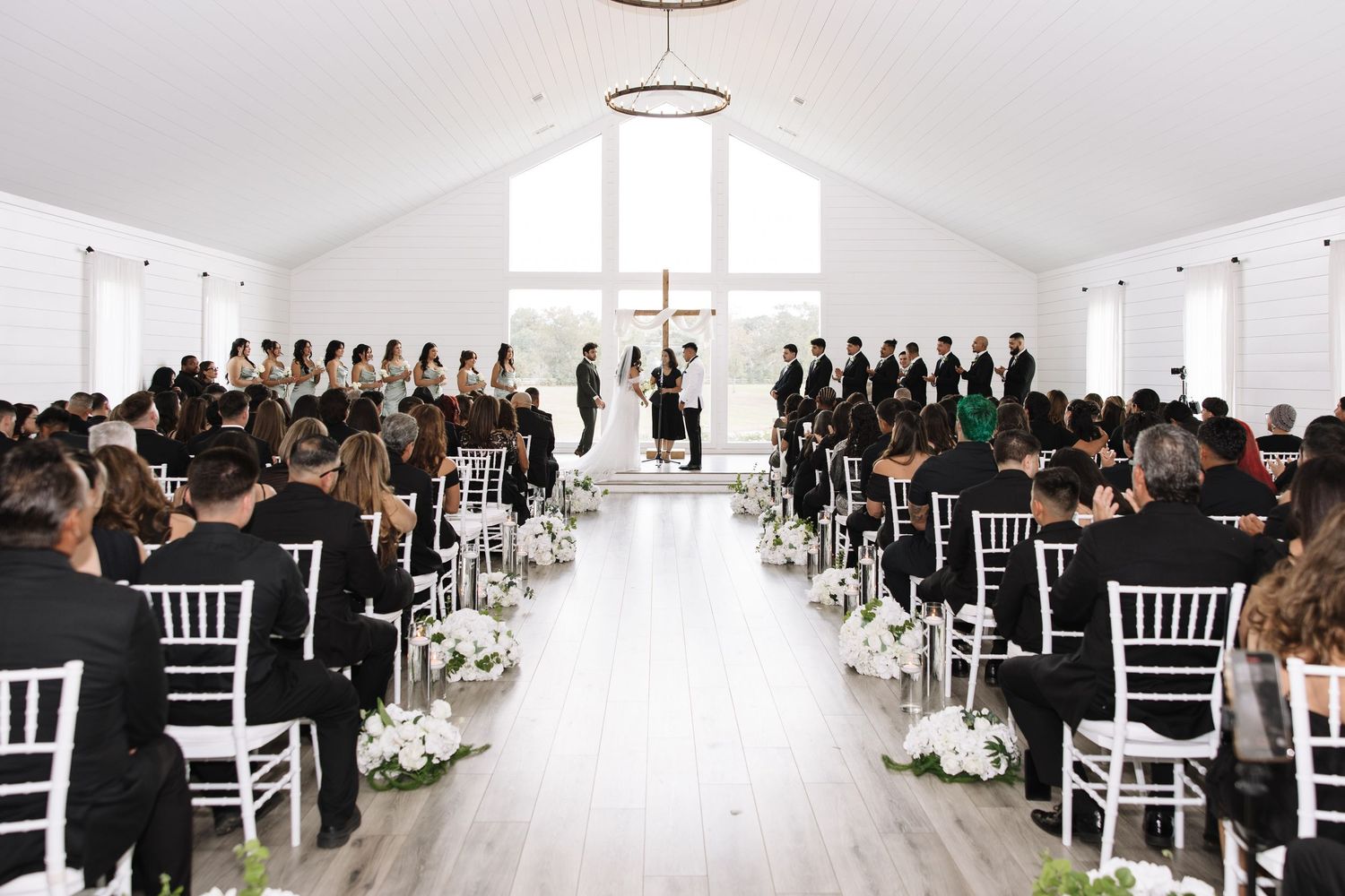 Wedding guests seated in white chairs during an indoor ceremony with elegant decor and lighting.