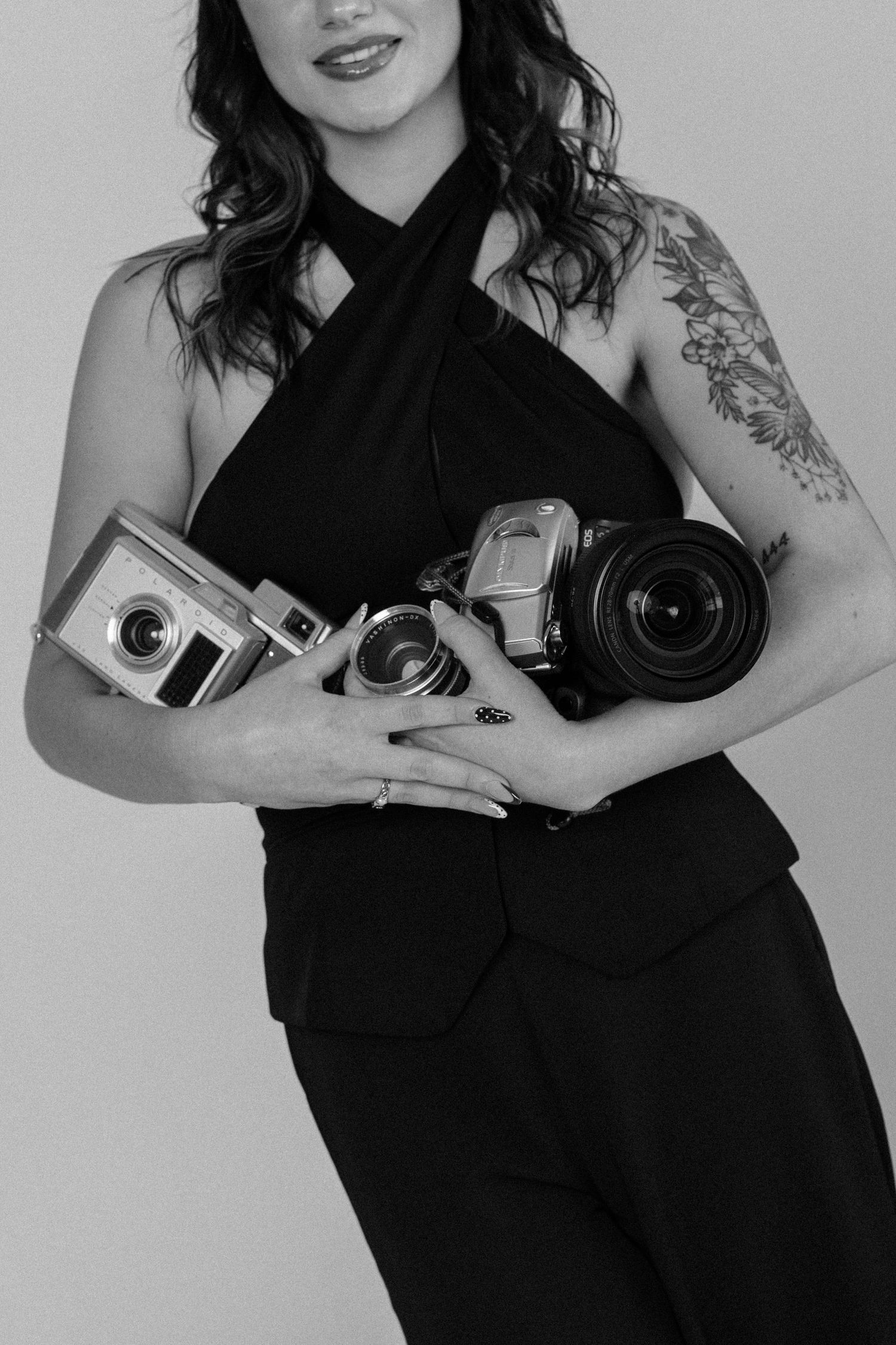 Black and white photo of a woman in black outfit holding multiple vintage and modern cameras, with a floral arm tattoo.