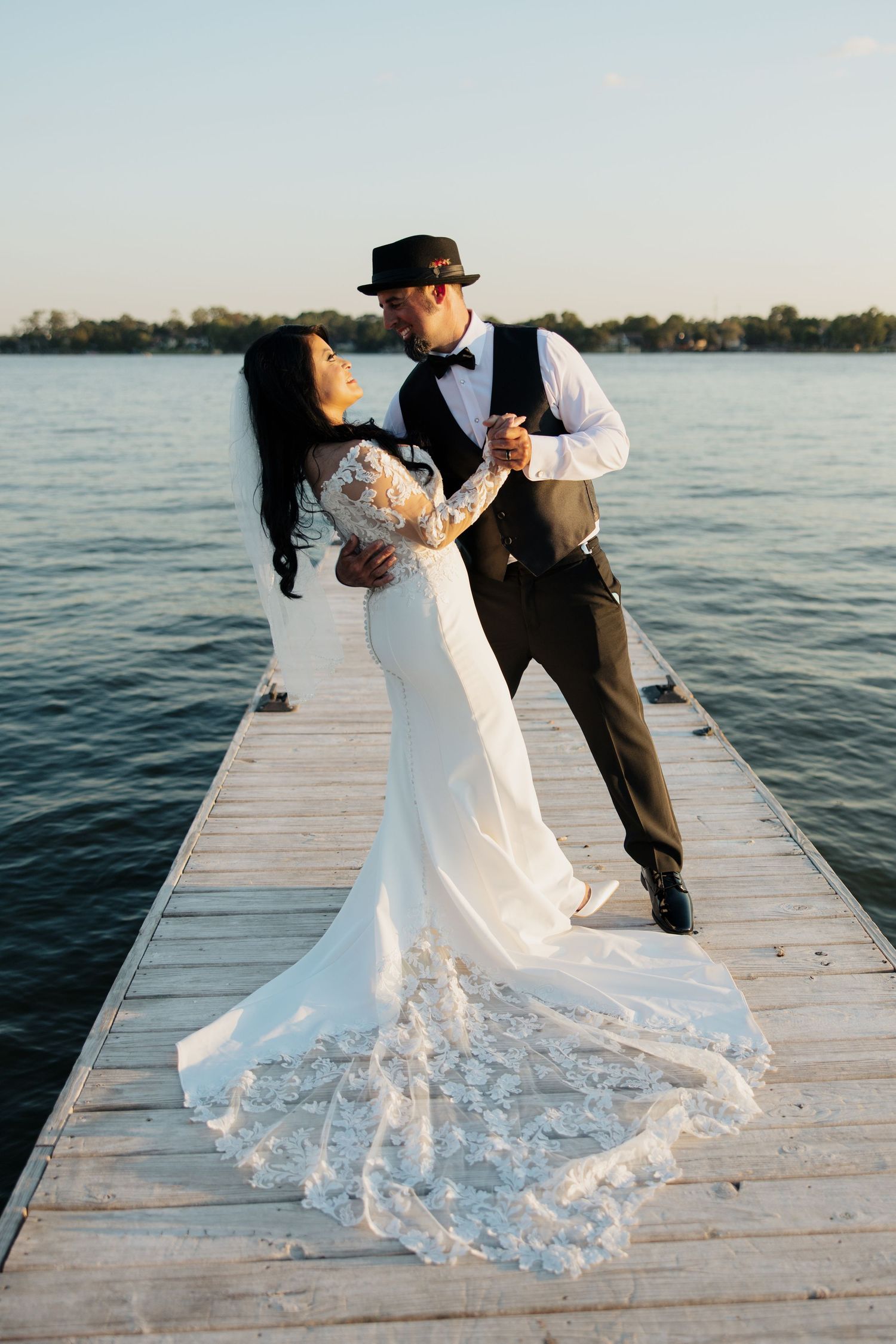 A couple dances romantically on a lakeside dock, with the bride's elegant white dress train flowing behind them.