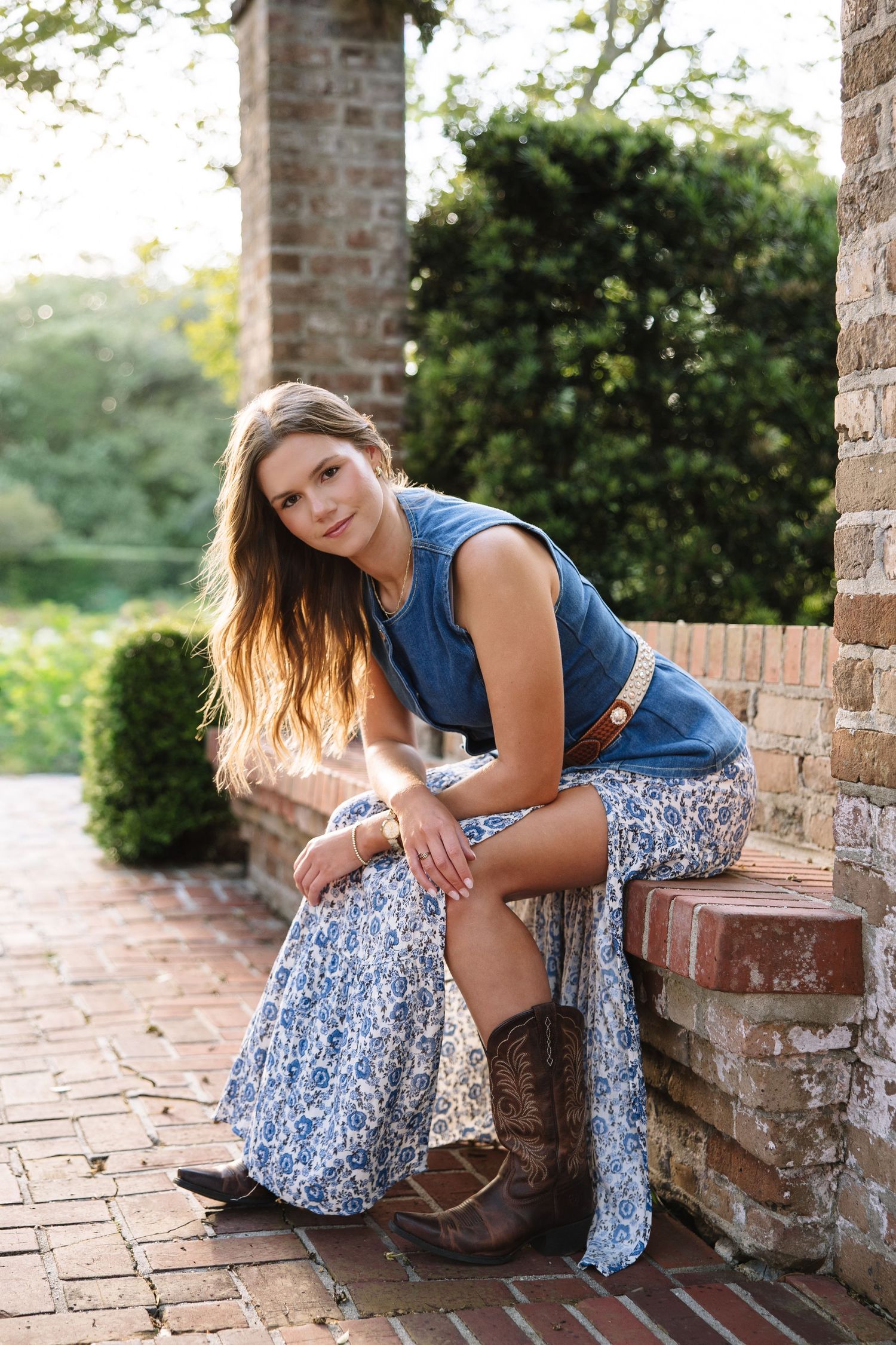 Young woman in denim top, floral maxi skirt, and cowboy boots sitting on brick steps outdoors.