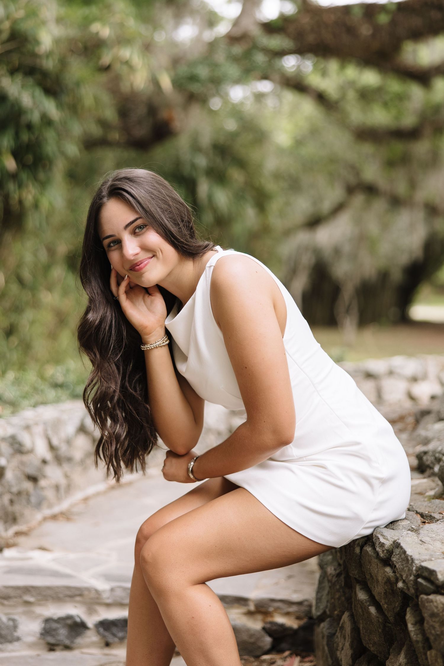 Young woman in white dress sitting on stone wall, smiling outdoors among lush green trees.