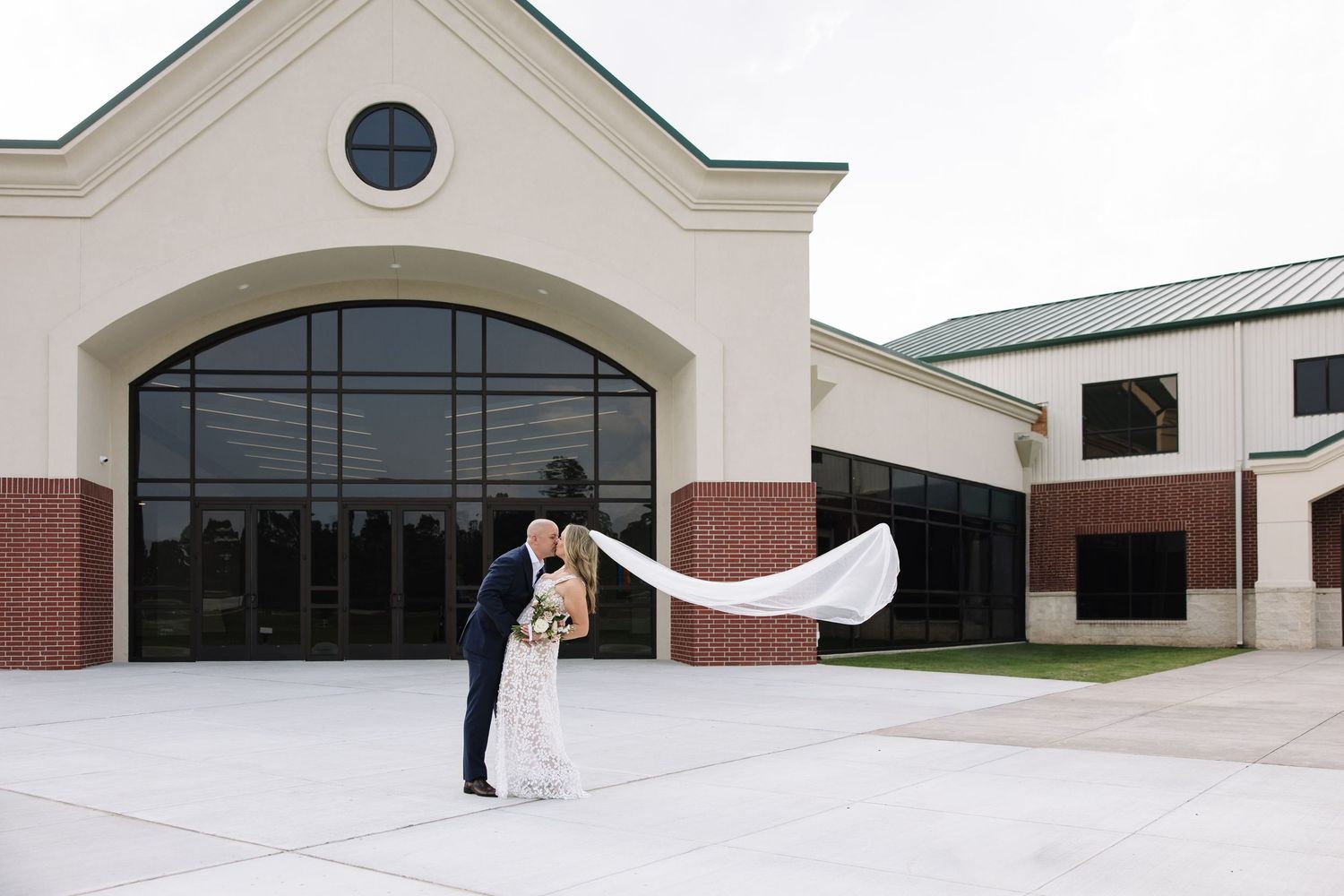 Bride and groom share a romantic dip kiss outside a modern wedding venue with veil flowing in the wind.