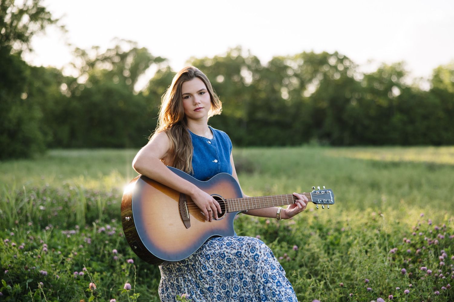 Young woman in blue top plays acoustic guitar while sitting in a lush green field at golden hour.