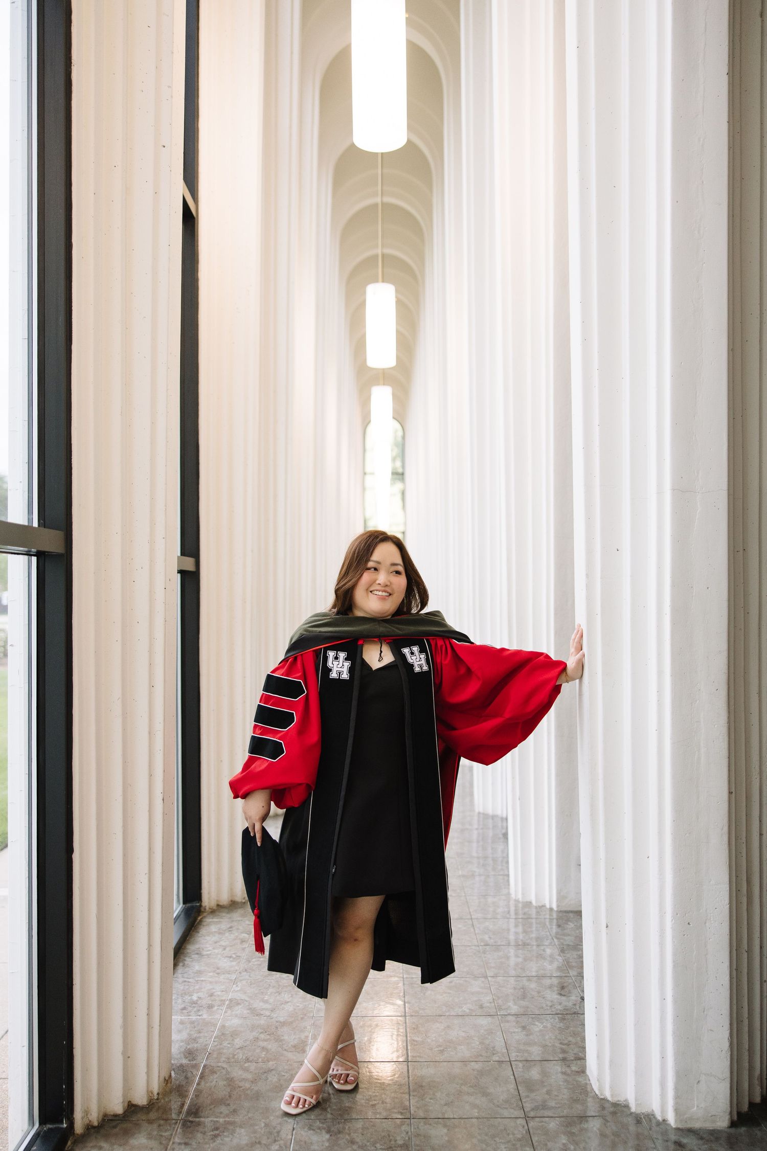 A woman in red and black doctoral graduation regalia smiles between white columns in an elegant hallway.