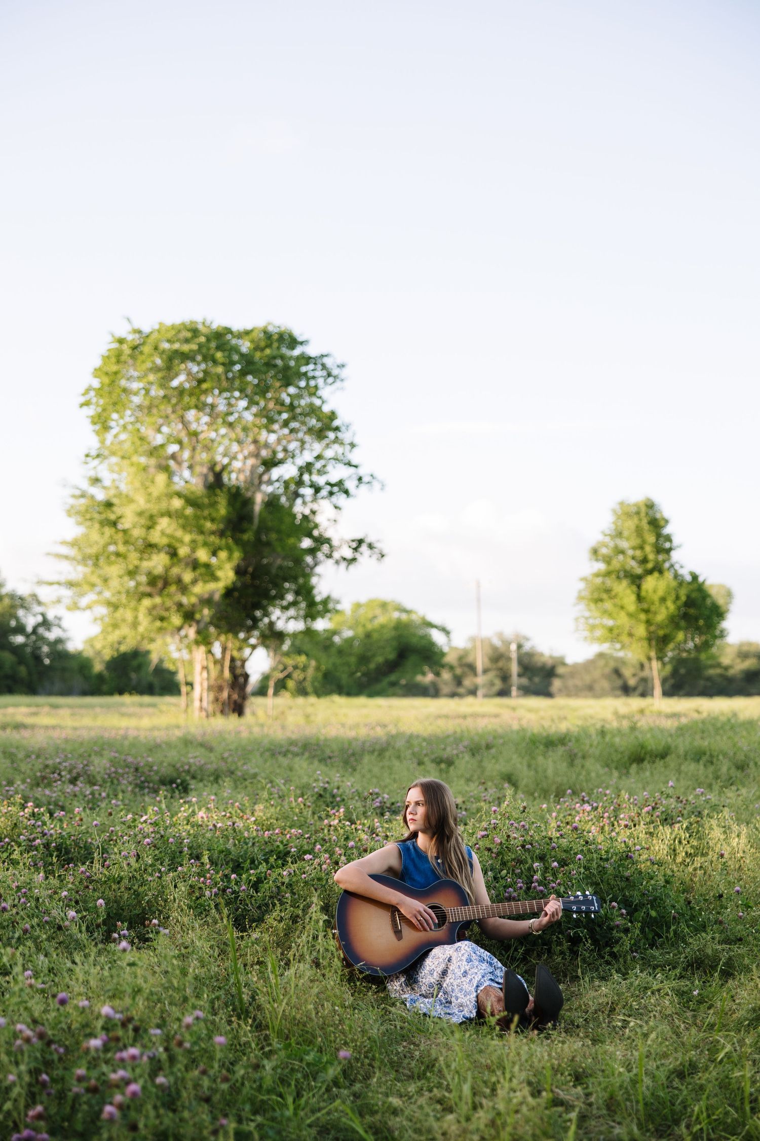 Young woman sitting in a wildflower field playing acoustic guitar surrounded by lush green trees.
