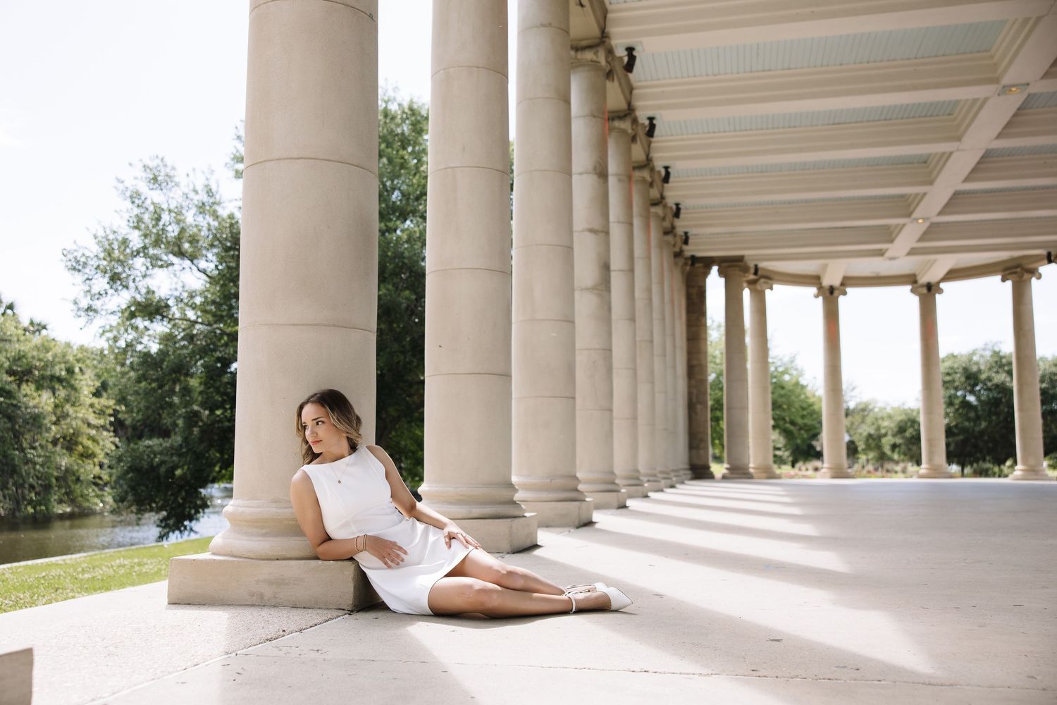 Woman in white dress sitting gracefully against marble columns in a sunlit classical colonnade park setting.