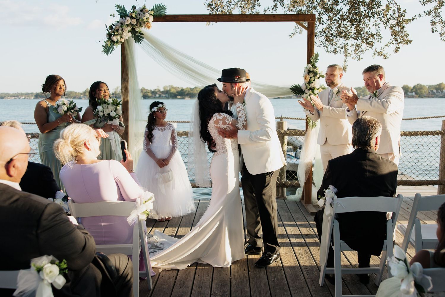 Romantic outdoor wedding ceremony on wooden deck overlooking waterfront with floral arch and seated guests.