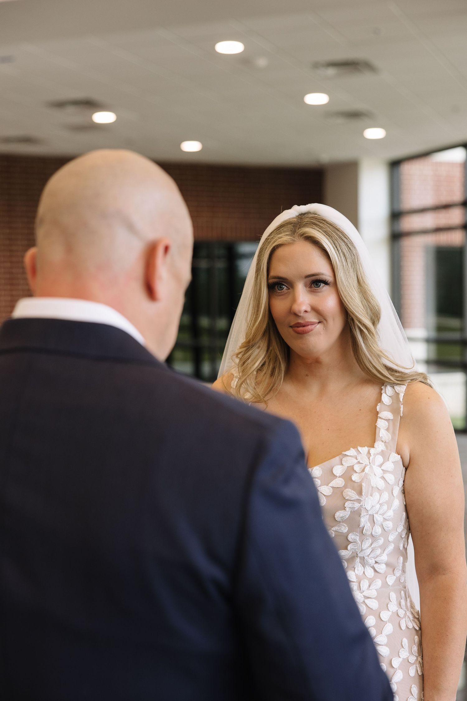 Bride in floral white gown gazes at groom in navy suit during wedding ceremony indoors.