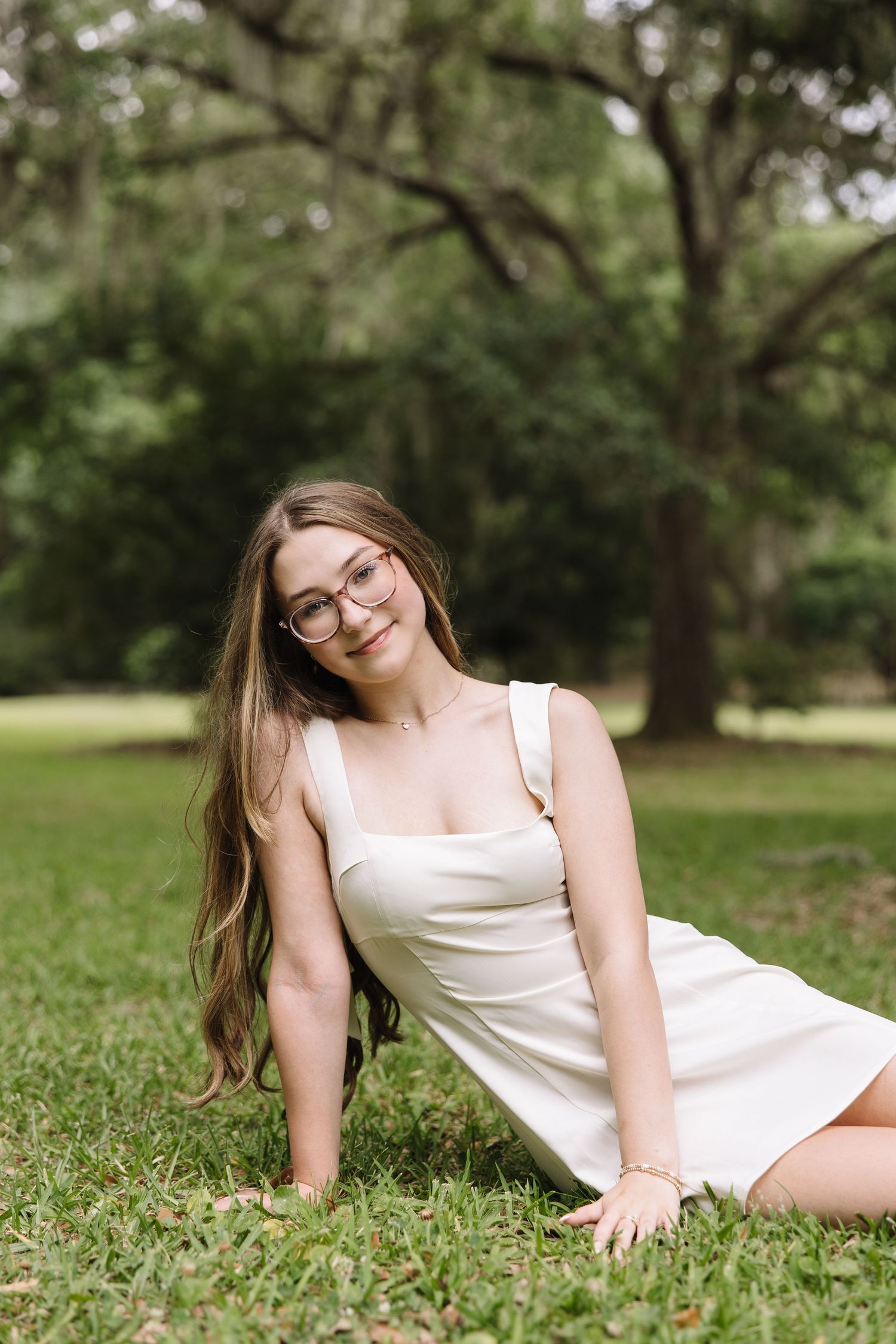 Young woman in white dress sitting on grass in park, smiling, wearing glasses, long hair, outdoor portrait.