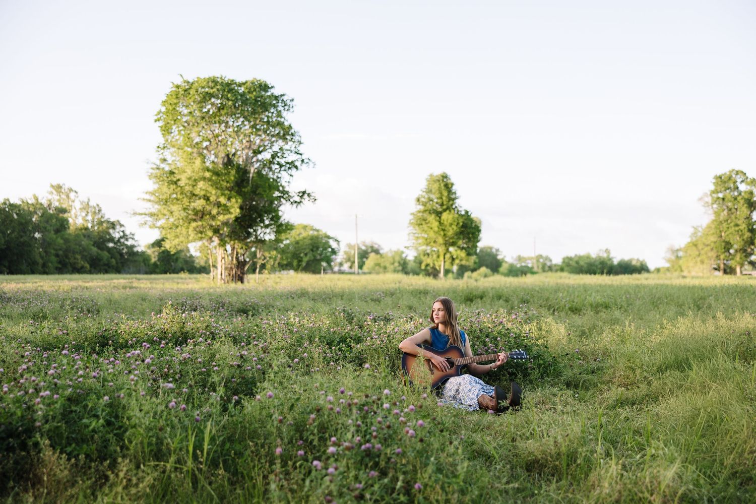 Woman playing guitar in a lush green meadow with wildflowers and trees at sunset.