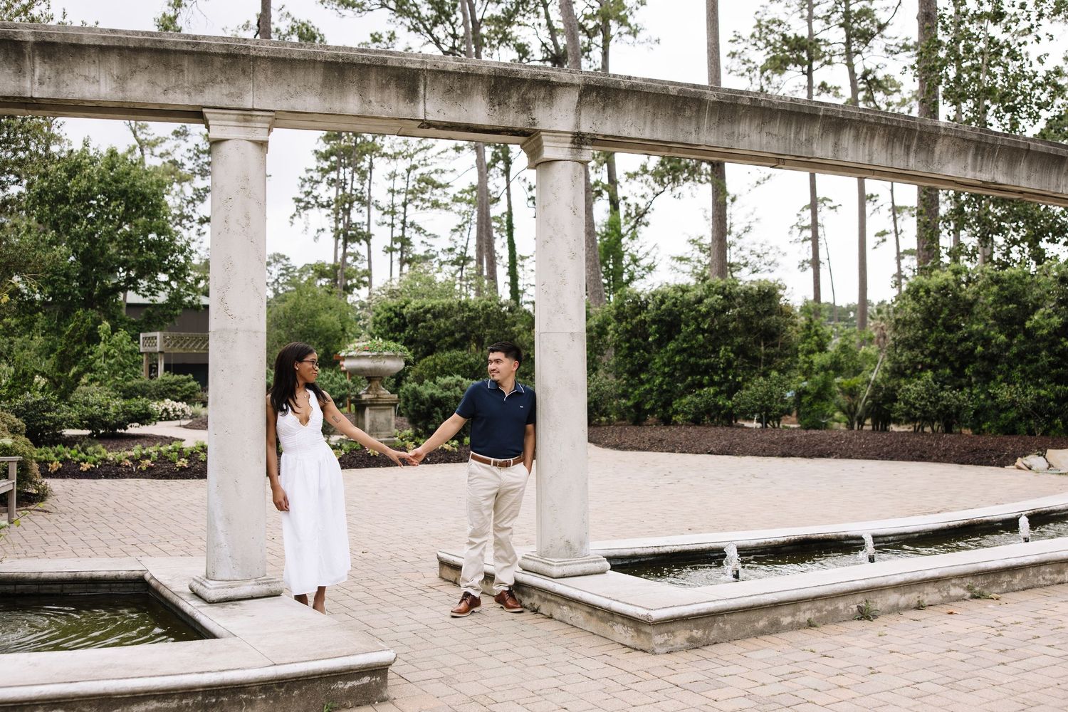 Couple holds hands in elegant garden with white columns, lush greenery, and brick pavement during outdoor engagement session.
