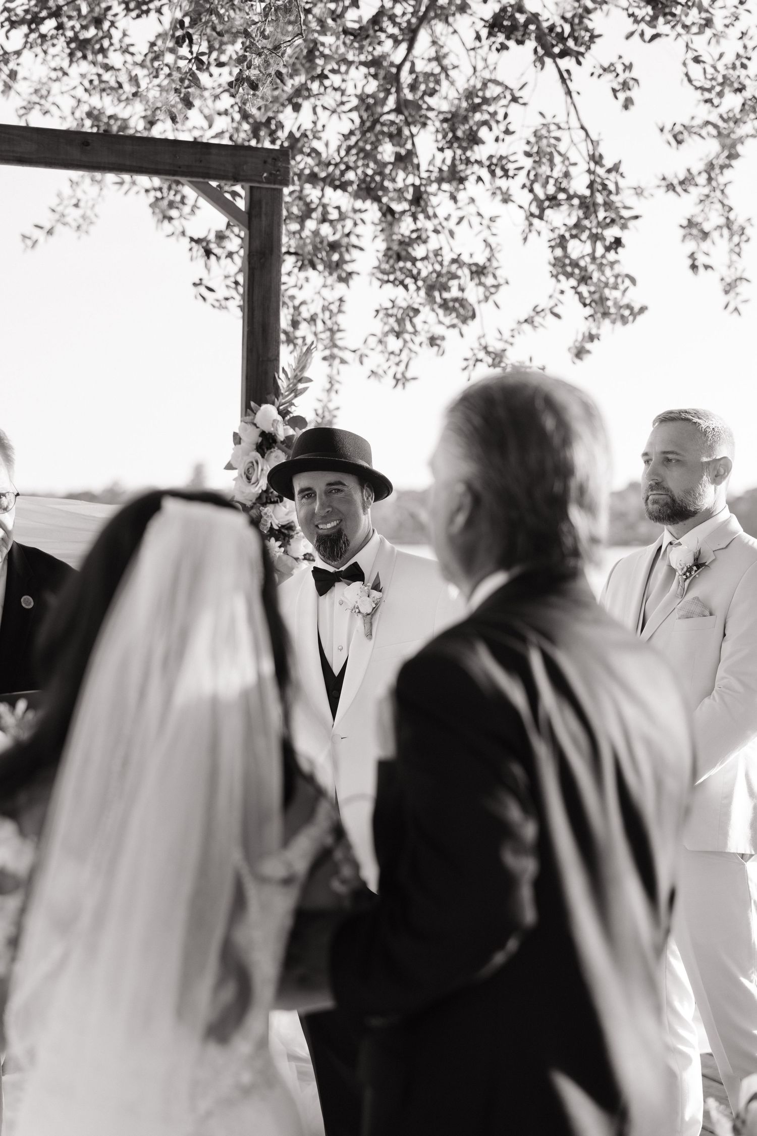 A black and white wedding ceremony moment captured under tree branches during an outdoor celebration.