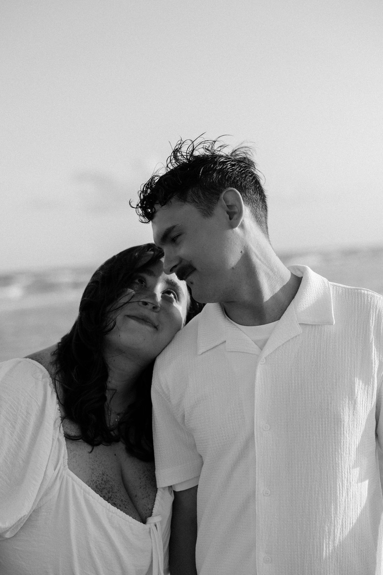 Black and white photo of a romantic couple sharing a tender moment on a beach, woman leaning on mans shoulder.