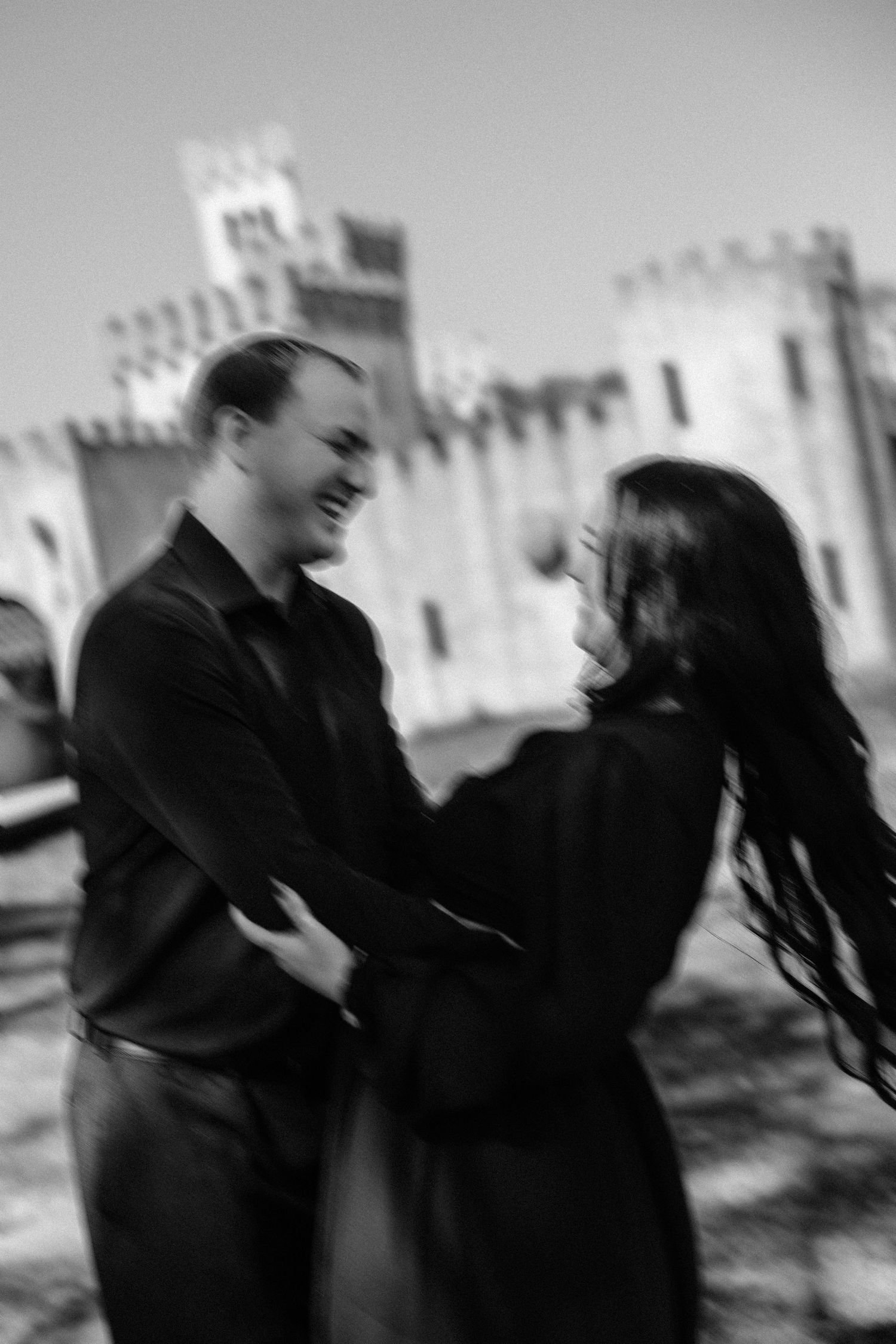 Black and white photo of a laughing couple dancing together in front of a historic castle building.