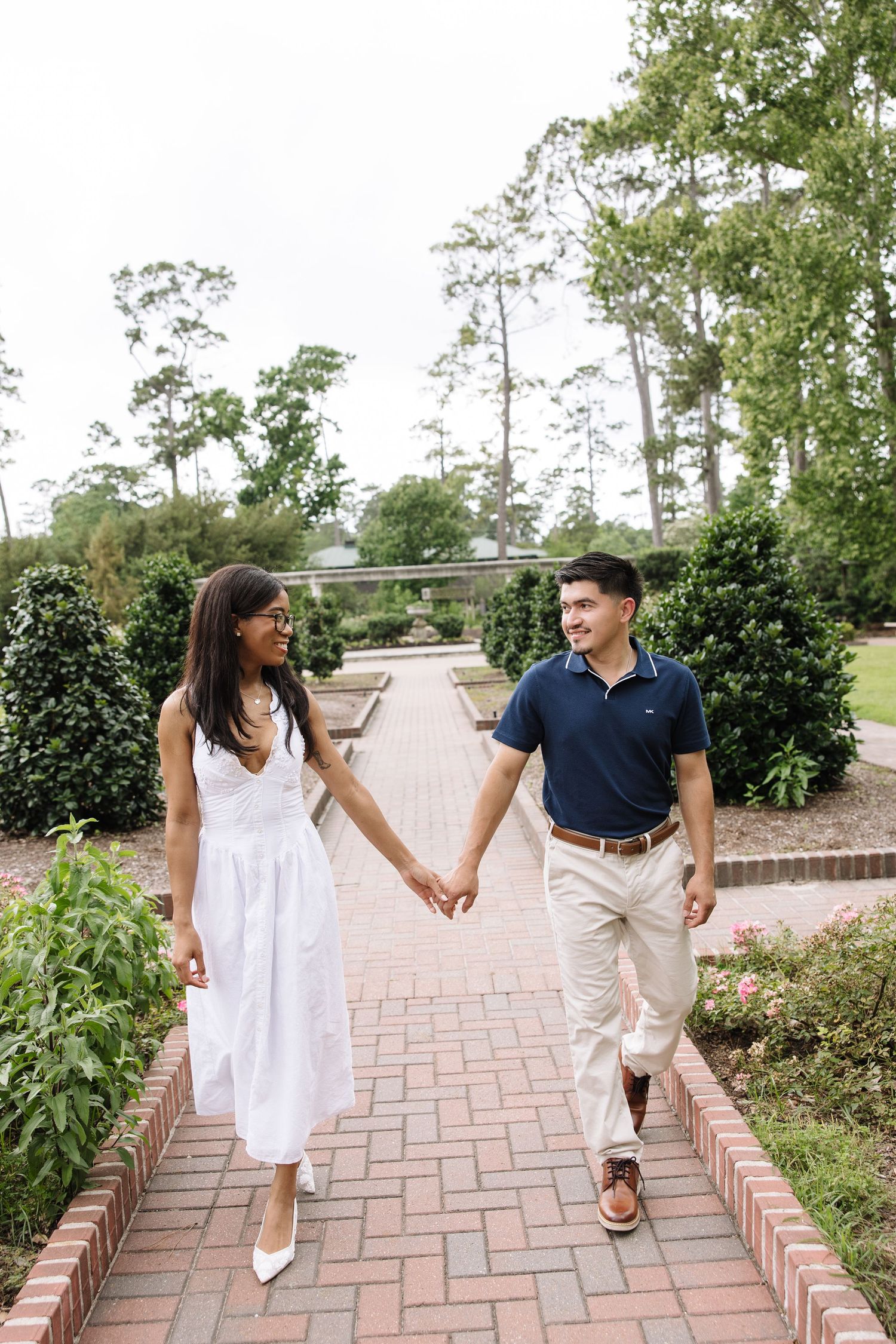 Couple holding hands and smiling at each other while walking along a brick garden path surrounded by lush greenery.