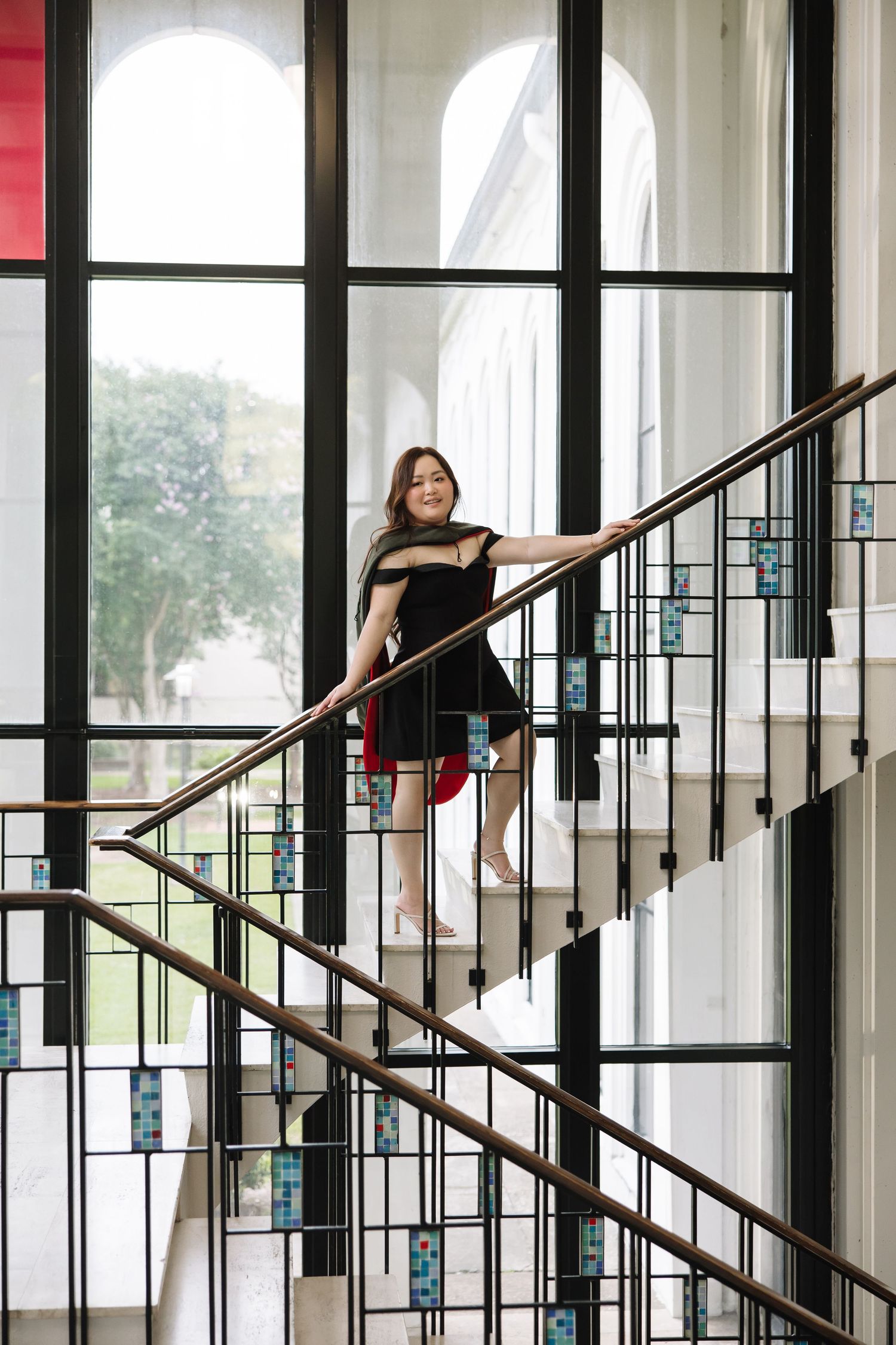 Woman in black dress posing on elegant staircase with decorative railings and arched windows in background.