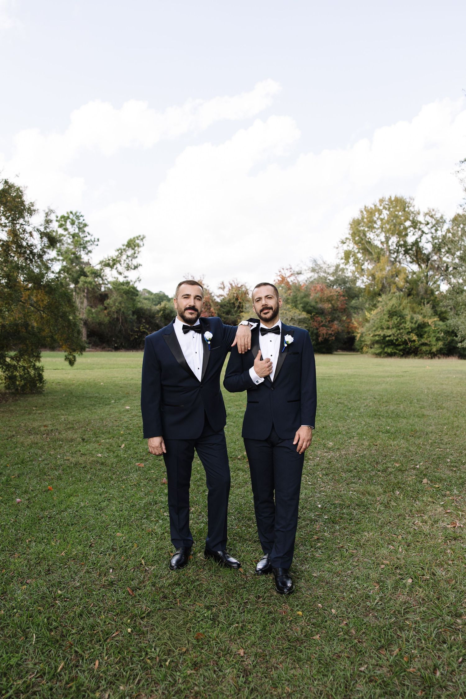 Groomsmen in black tuxedos pose together on a grassy field during an outdoor wedding photoshoot.