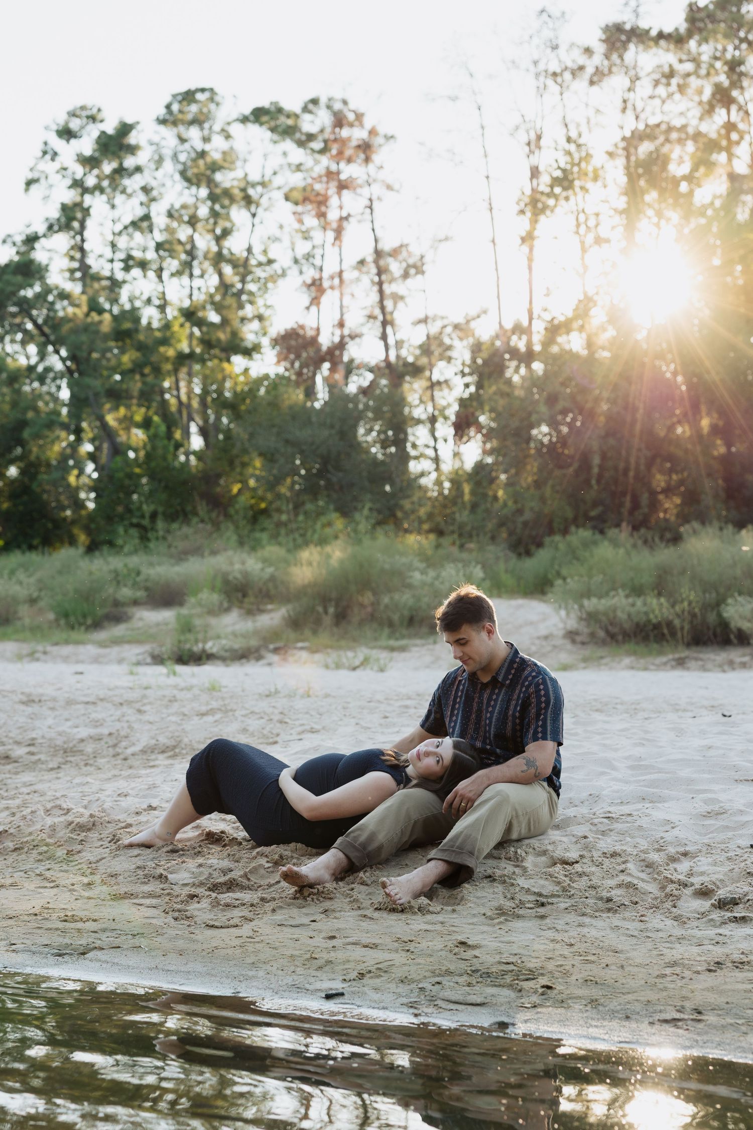 Couple relaxing together on a sandy beach at sunset with golden sunlight filtering through trees.