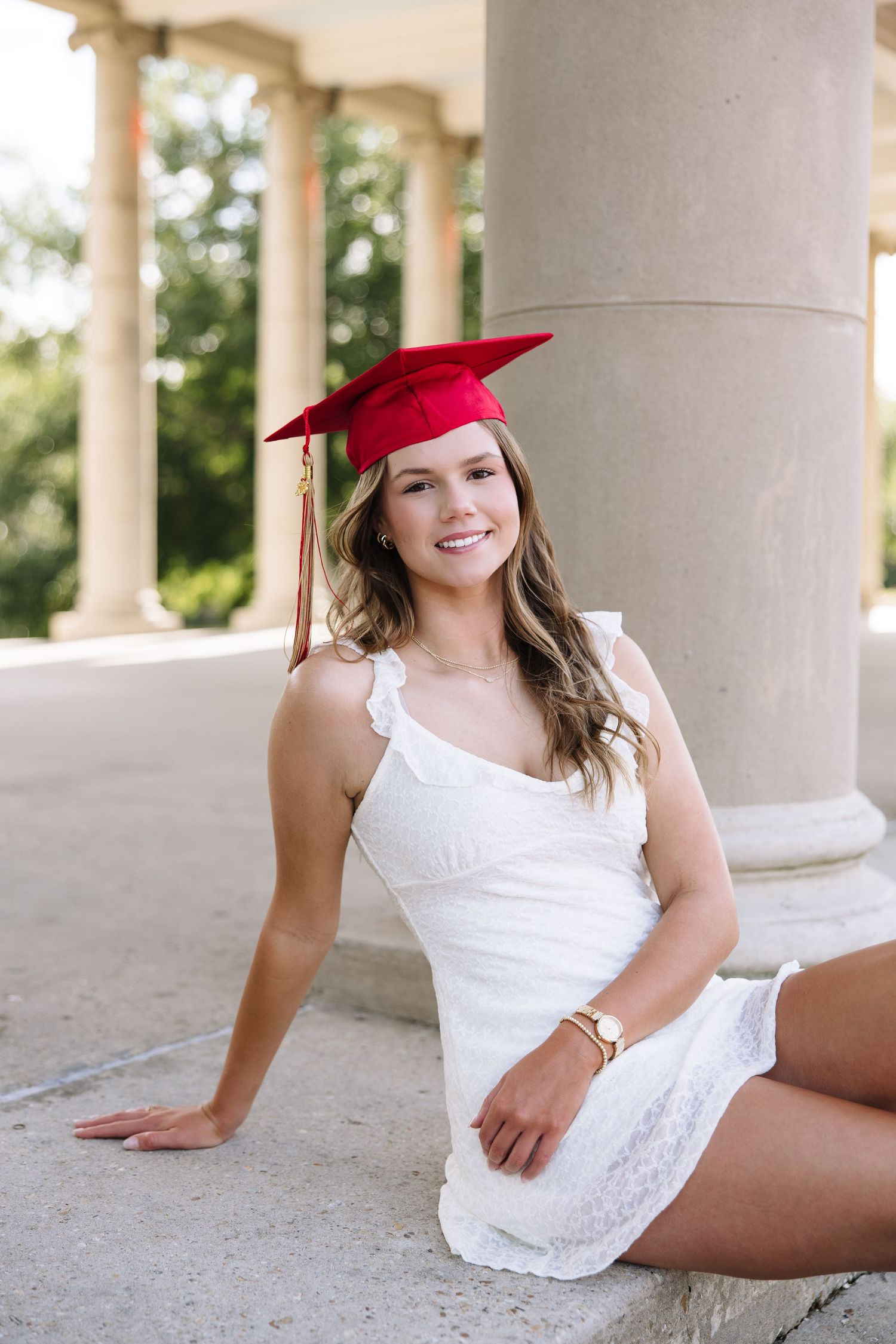 Young woman in white dress and red graduation cap sits smiling by stone columns outdoors.