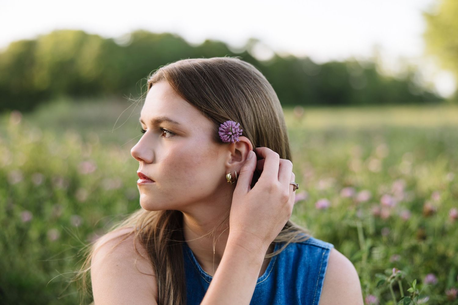 Young woman with freckles placing a purple wildflower in her hair in a sunlit meadow field.