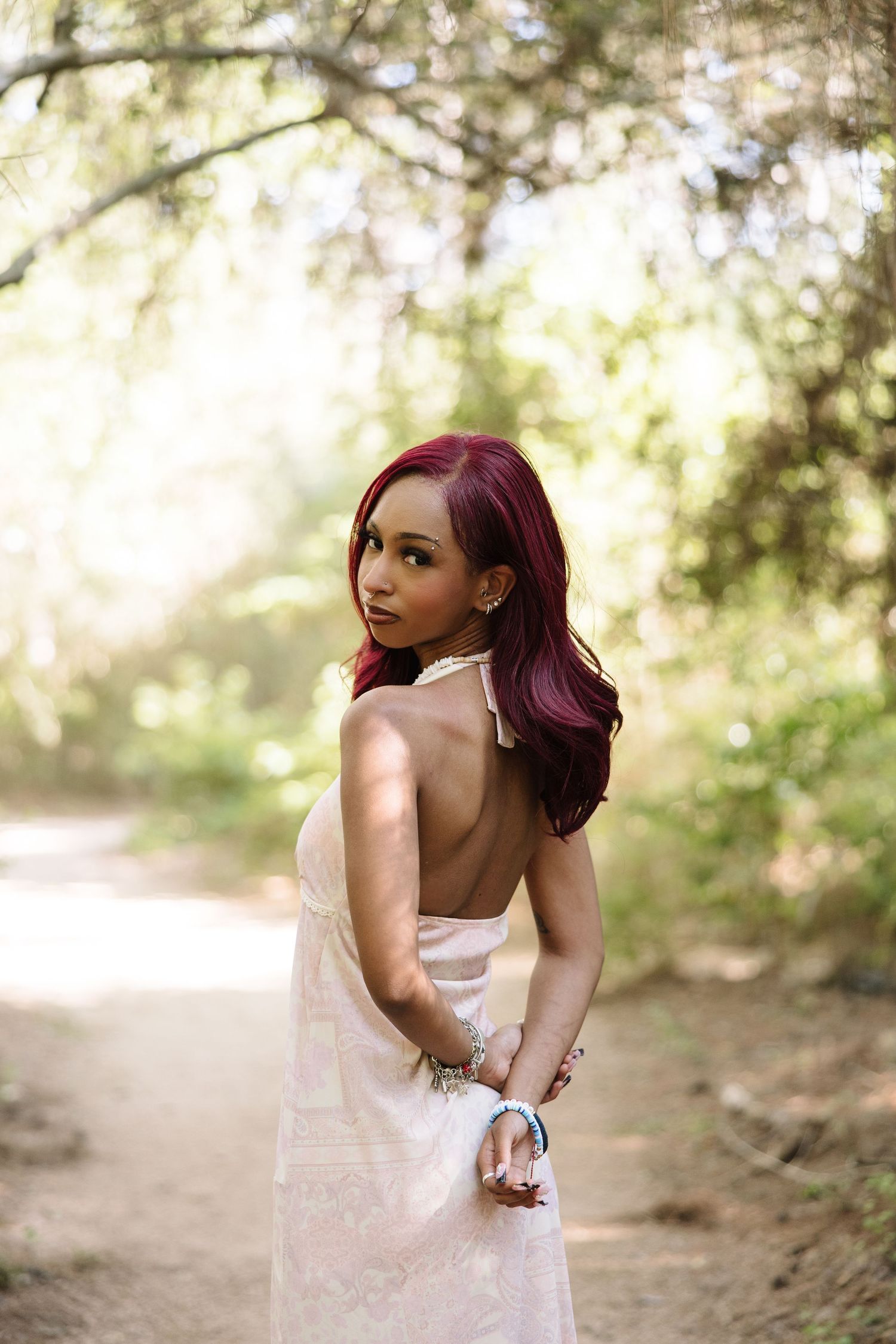 Woman with red hair in backless white dress poses on sunlit forest path.