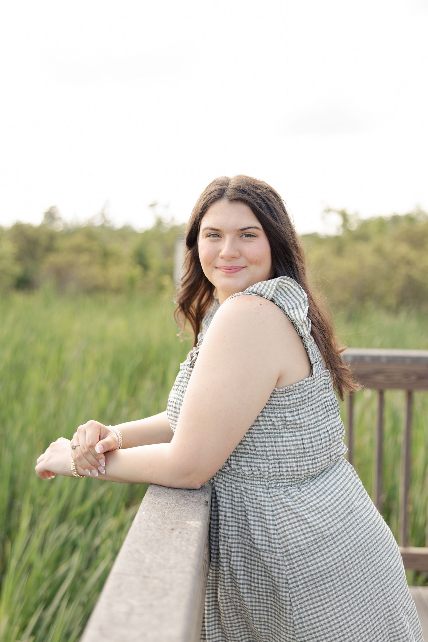 Woman in a gingham dress poses against a wooden railing with tall grass in the background.