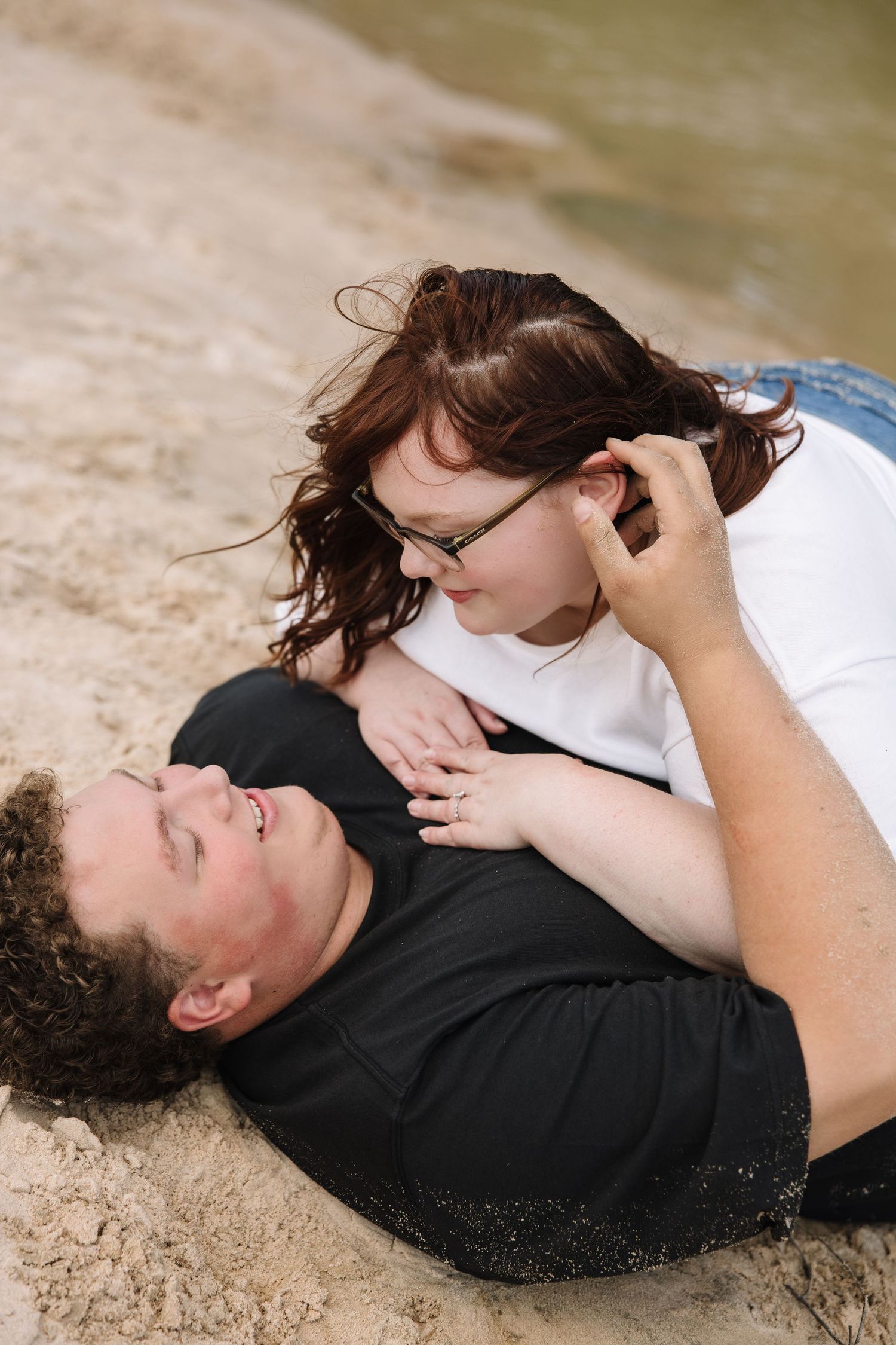 Couple relaxing on sandy beach, woman in white top leaning over smiling man in black shirt lying in sand.