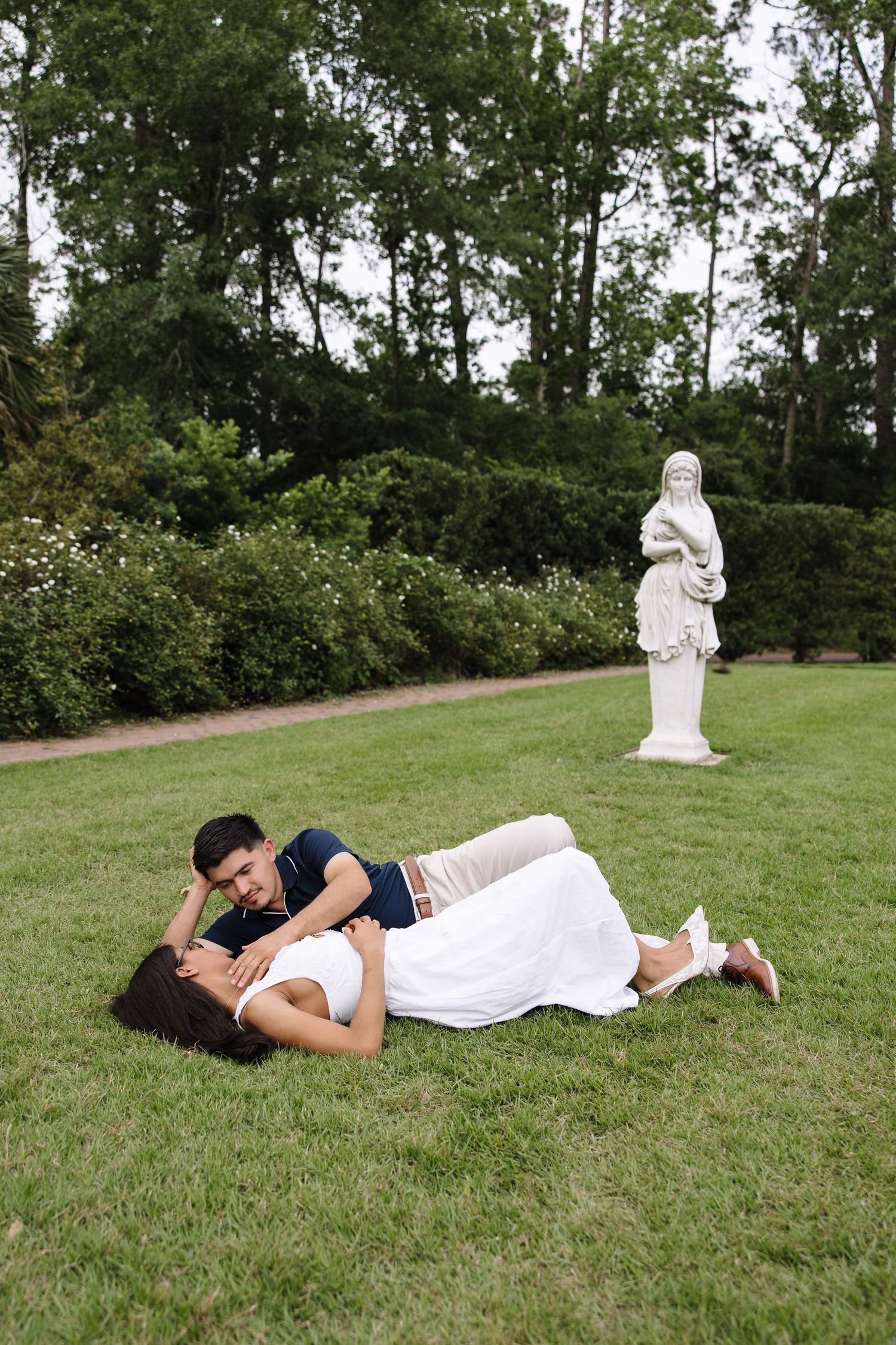Romantic couple lying on green grass in a garden with a white classical statue and lush trees in the background.