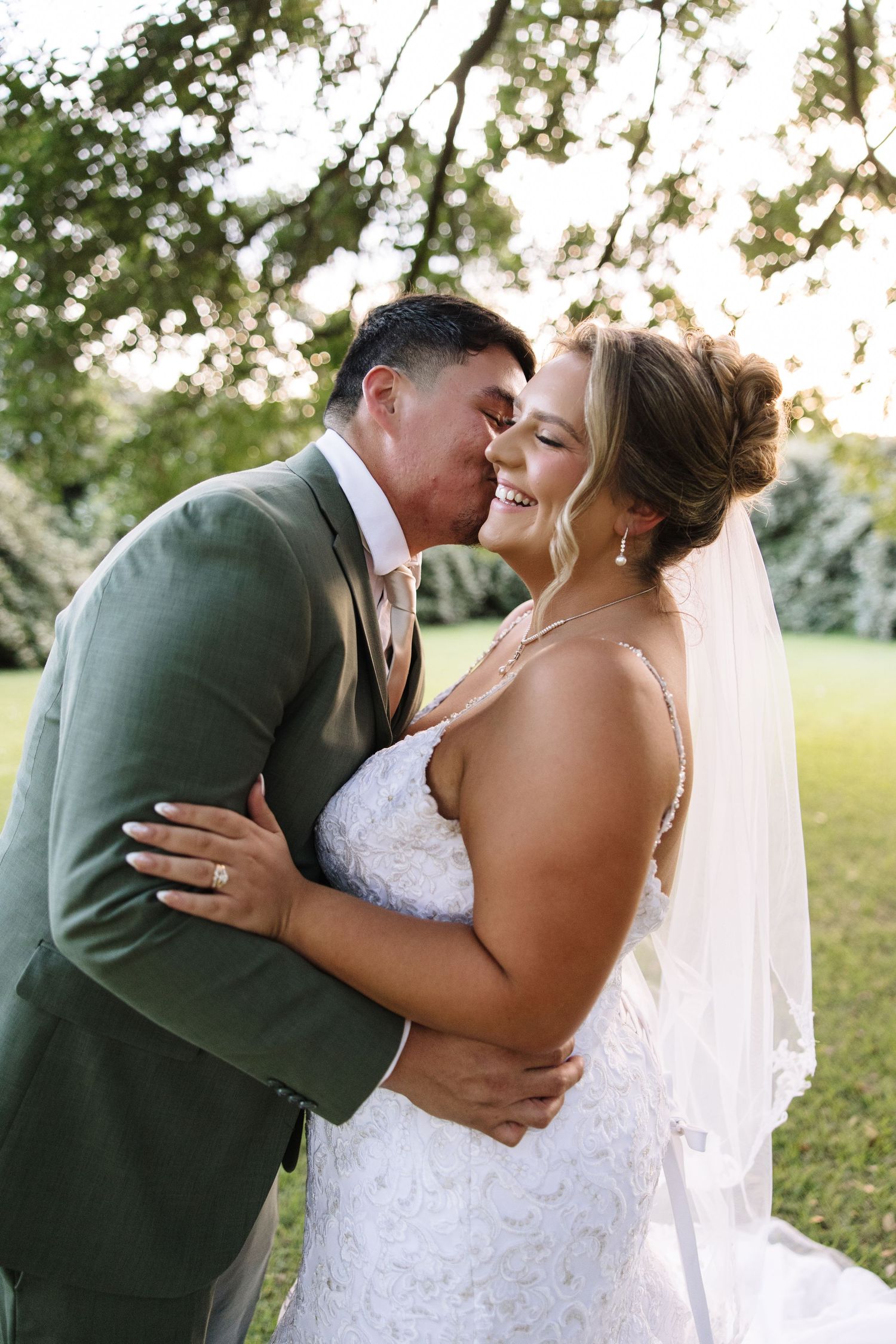 Bride and groom share a romantic kiss outdoors, groom in green suit, bride in lace gown with veil.