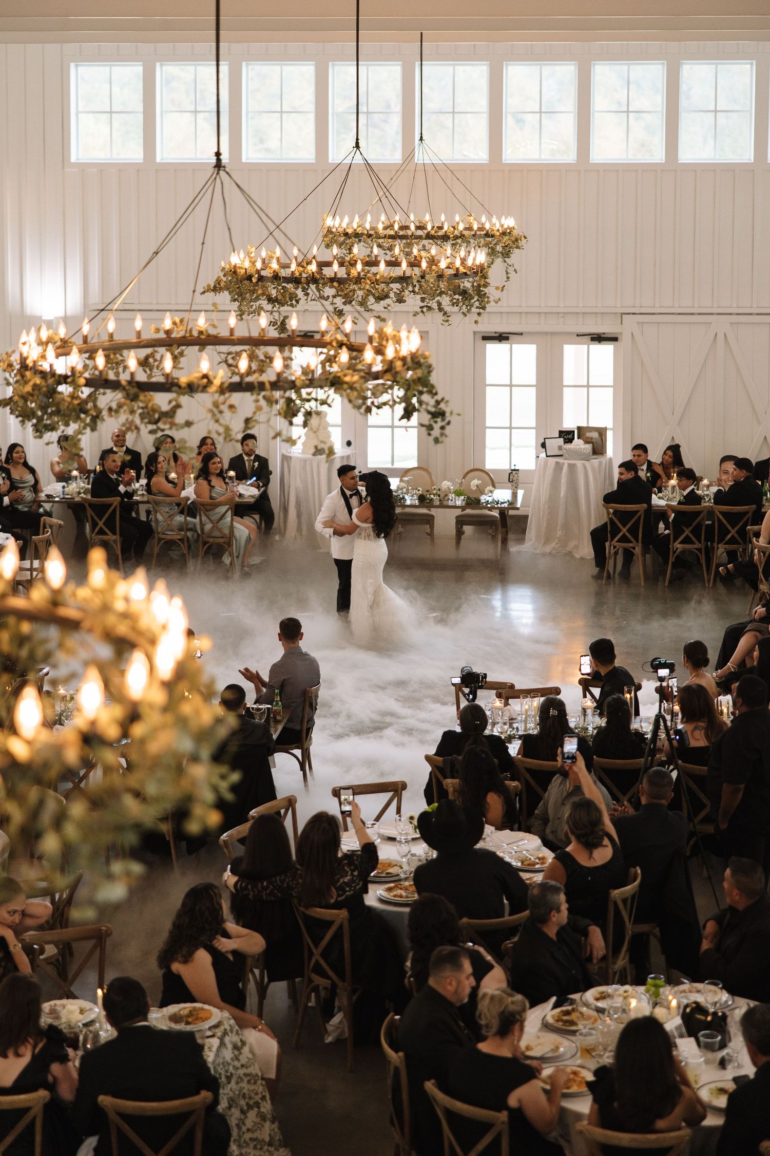 Wedding reception guests seated at tables under elegant crystal chandeliers in a bright white modern venue.