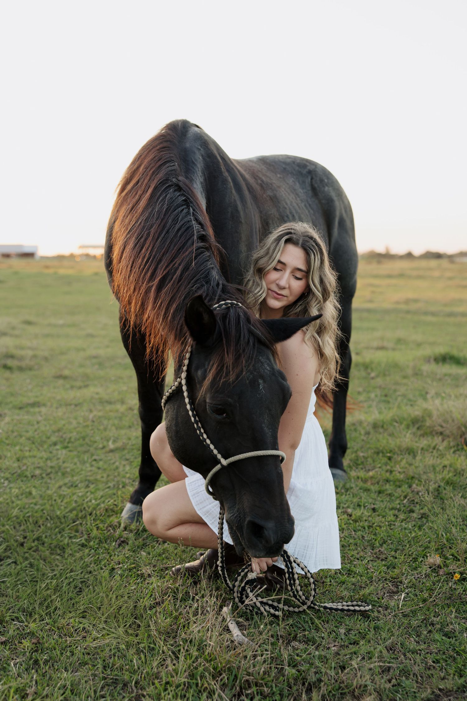 A person in a white dress shares a tender moment with a black horse grazing in a grassy field at sunset.