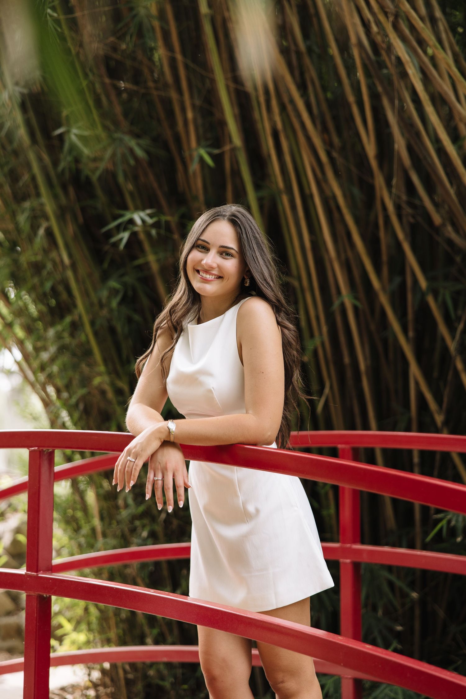 Young woman in white dress smiling on red bridge with bamboo trees in background.