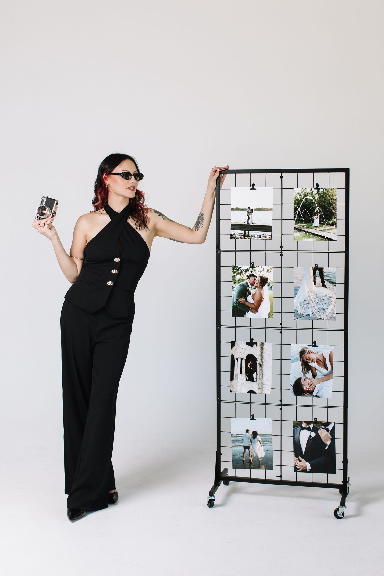 A stylish woman in black jumpsuit and sunglasses poses beside a grid display of wedding photos, holding a camera.