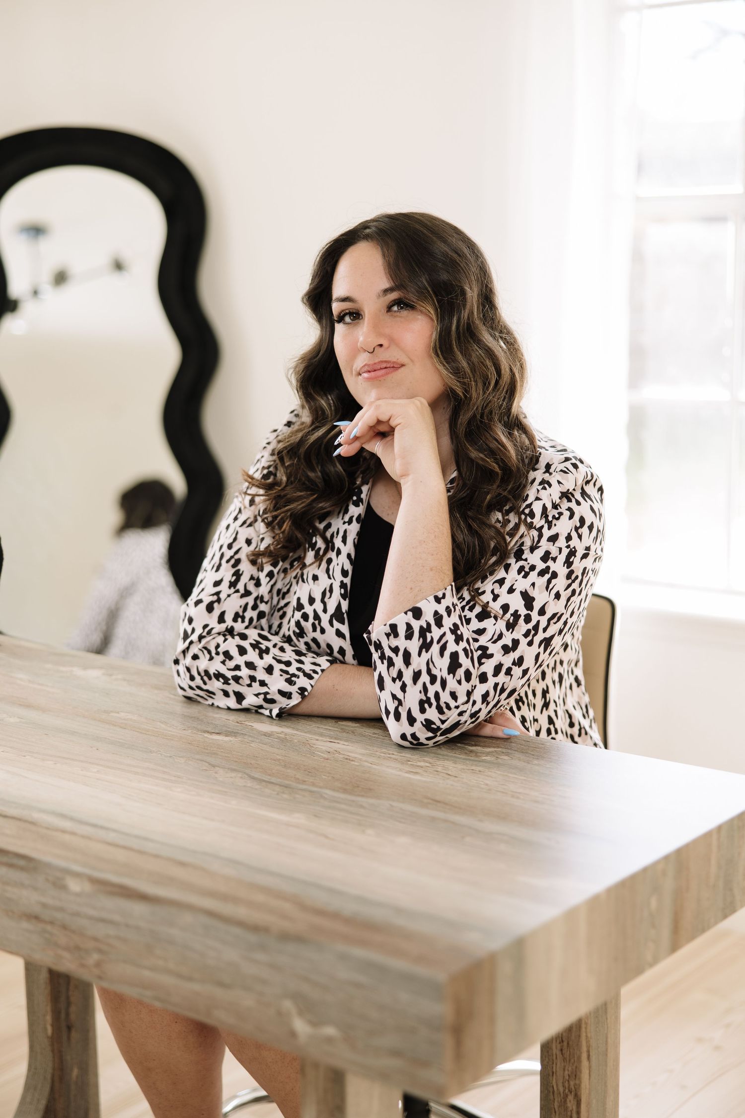 A person in a leopard print blouse leans on a wooden table in a bright white room.