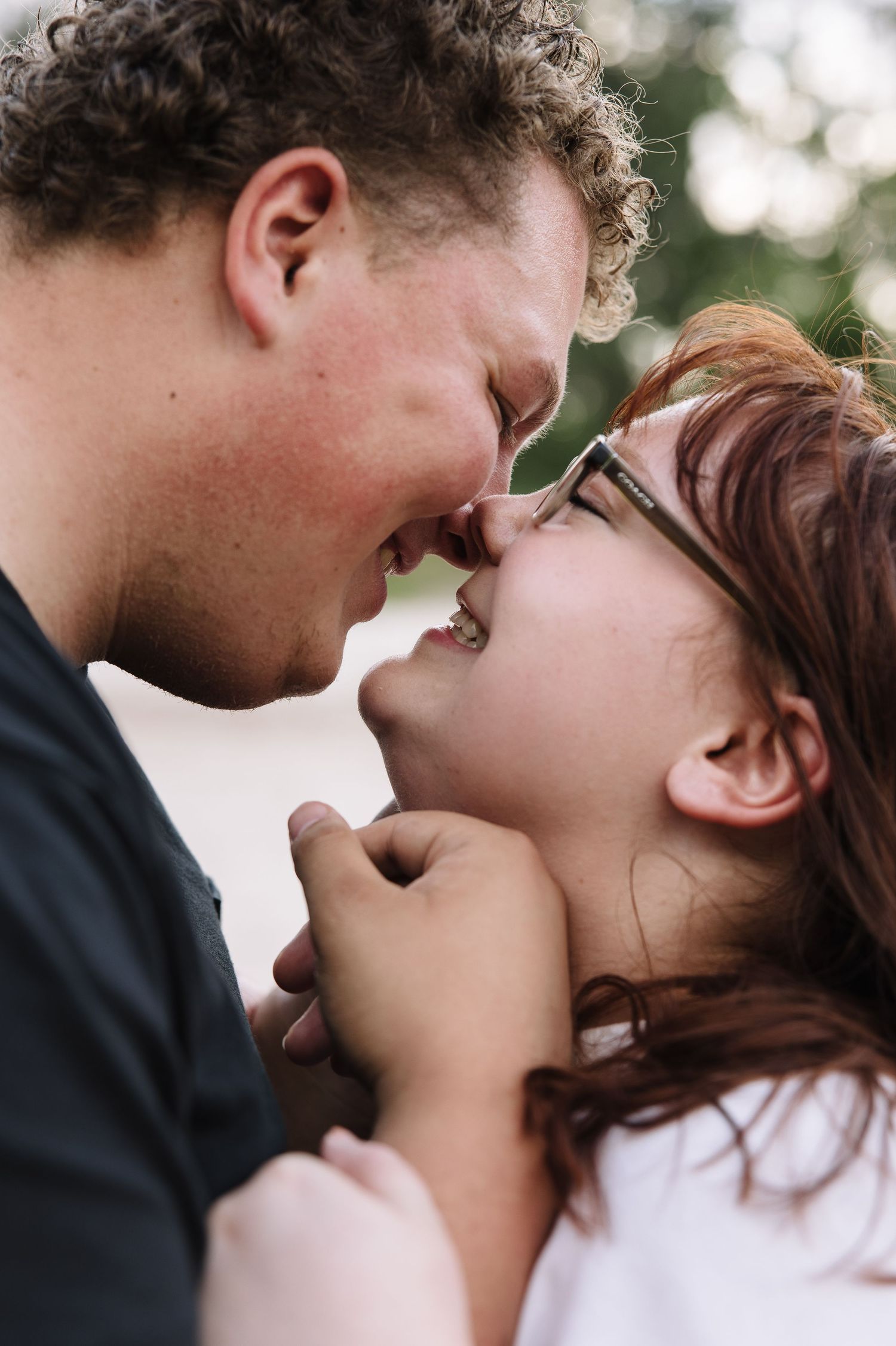 A couple shares a tender romantic moment, foreheads touching and hands clasped, smiling at each other outdoors.