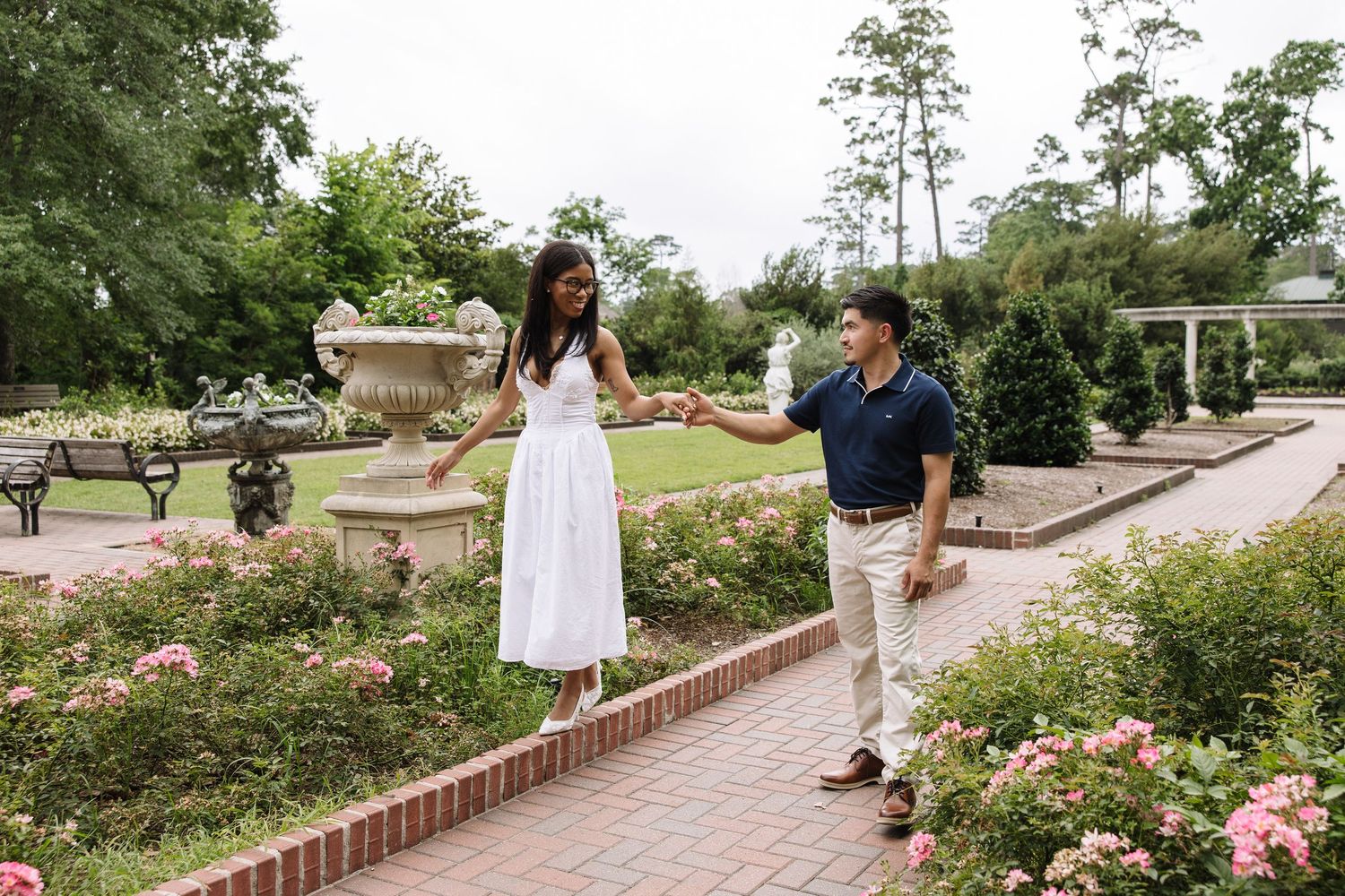 Couple holding hands and walking through a lush garden with pink roses and stone fountains on a brick pathway.