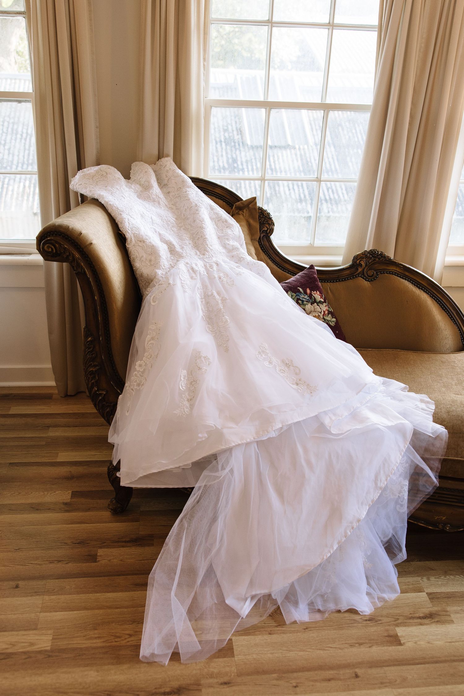White lace wedding gown draped over an ornate vintage chaise lounge near a sunlit window.
