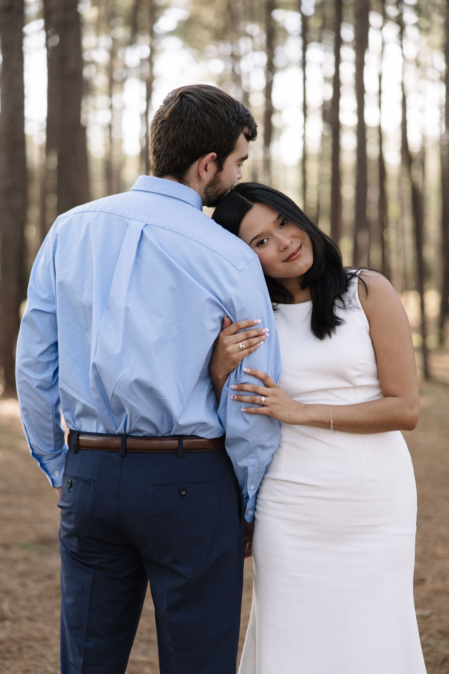 A romantic couple embraces in a forest setting, with the woman wearing white and the man in a blue shirt and navy pants.