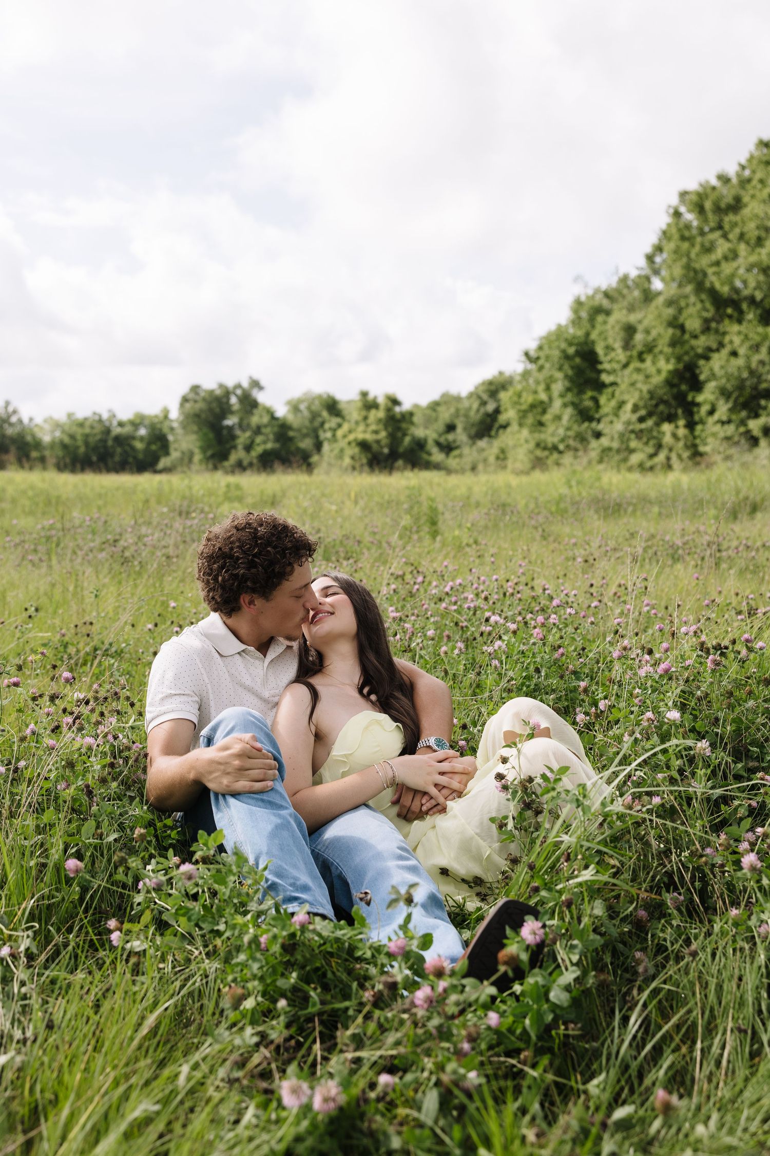 Romantic couple sitting in a wildflower meadow, sharing a tender kiss surrounded by green trees.