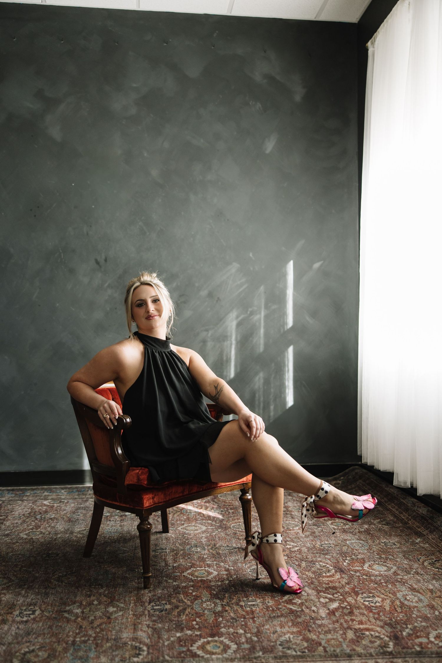 Artistic boudoir photo series showing figure in black dress reclining on vintage chair against dark wall.