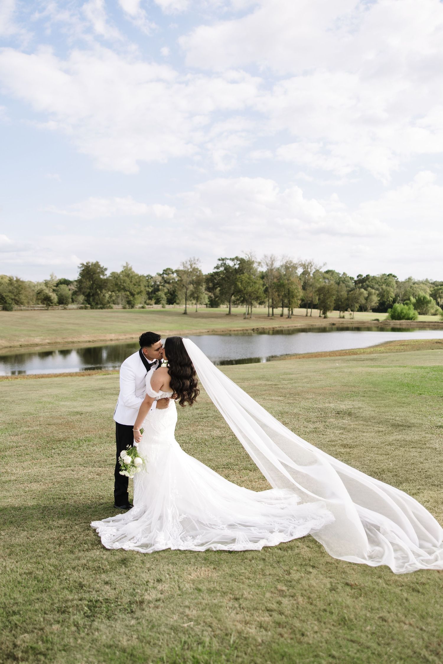 A romantic wedding couple poses in a grassy field with a flowing long veil creating a dramatic effect near a lake.