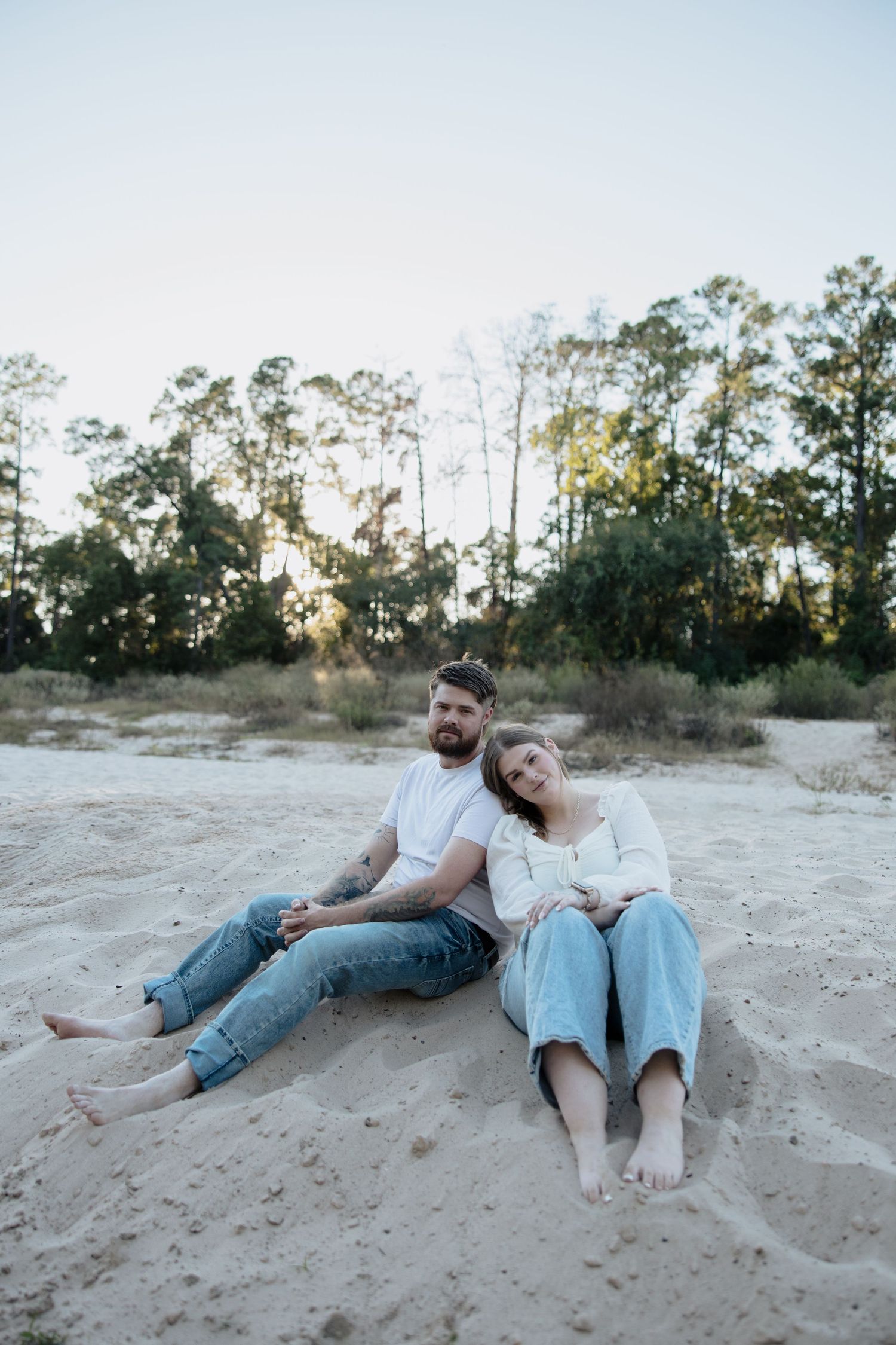 A couple sits together on a sandy beach at sunset with tall pine trees in the background.
