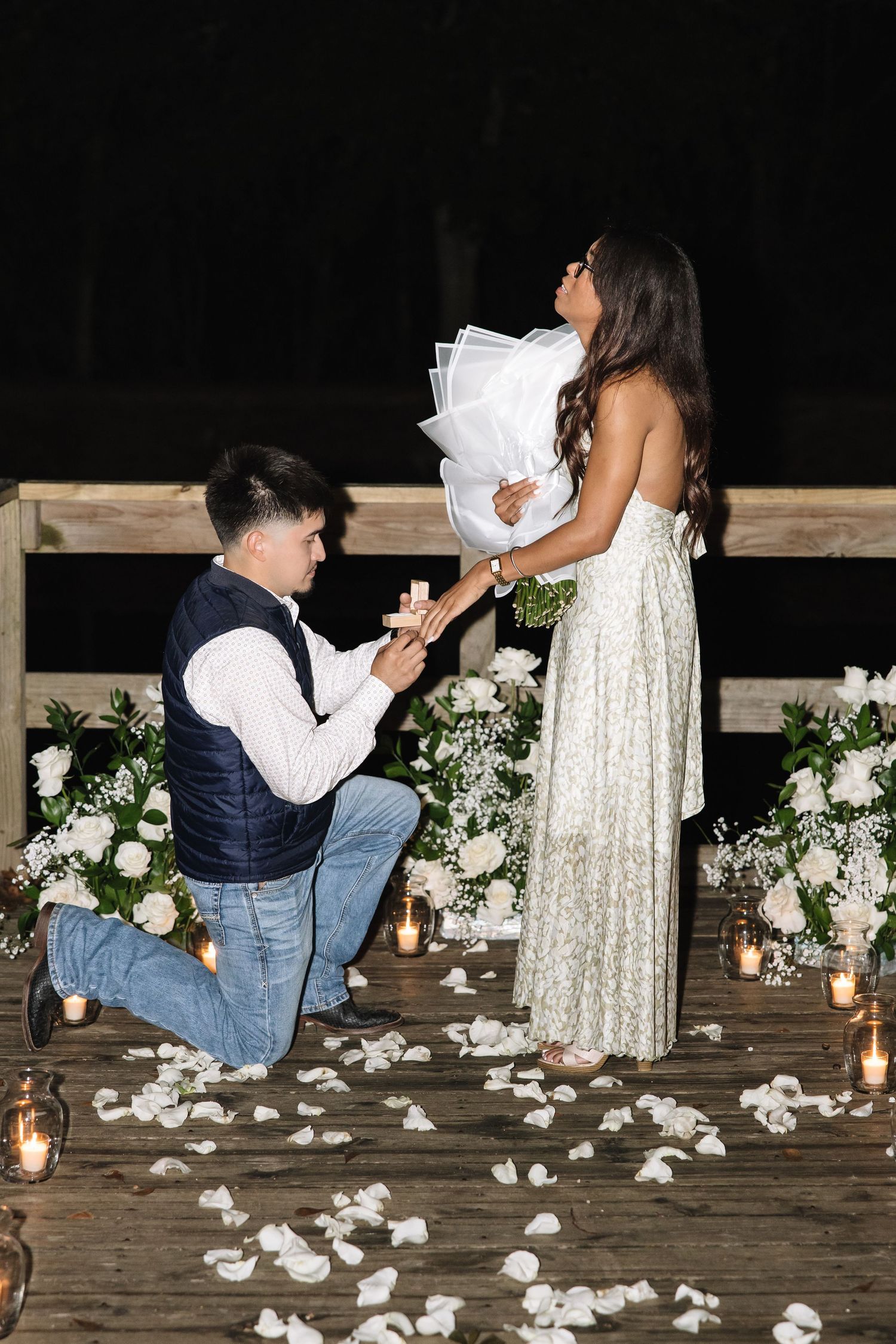 A romantic marriage proposal sequence on a wooden deck decorated with white flowers and candles at night.