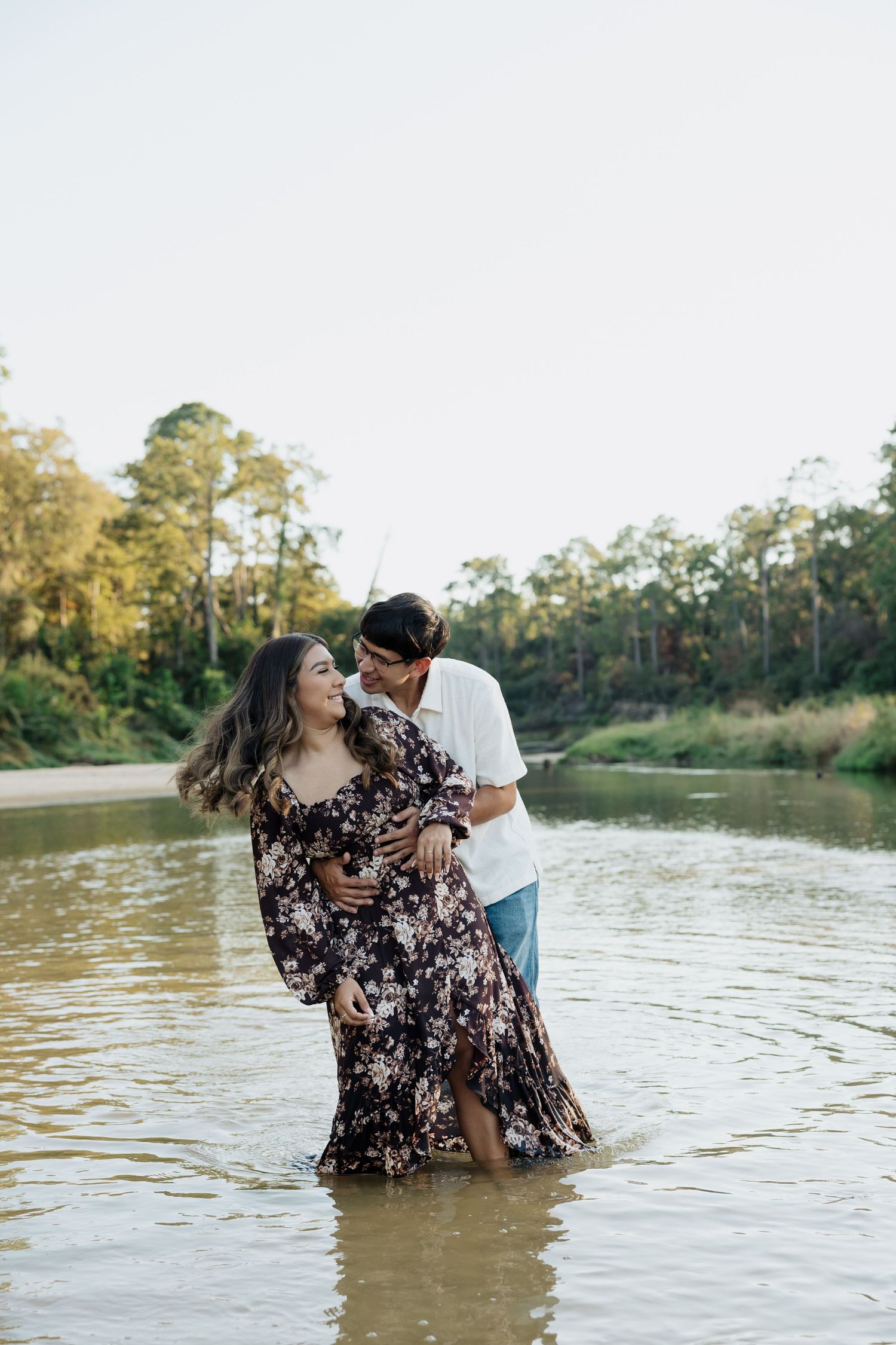 A romantic couple embraces while standing in a lake with trees in the background during golden hour.